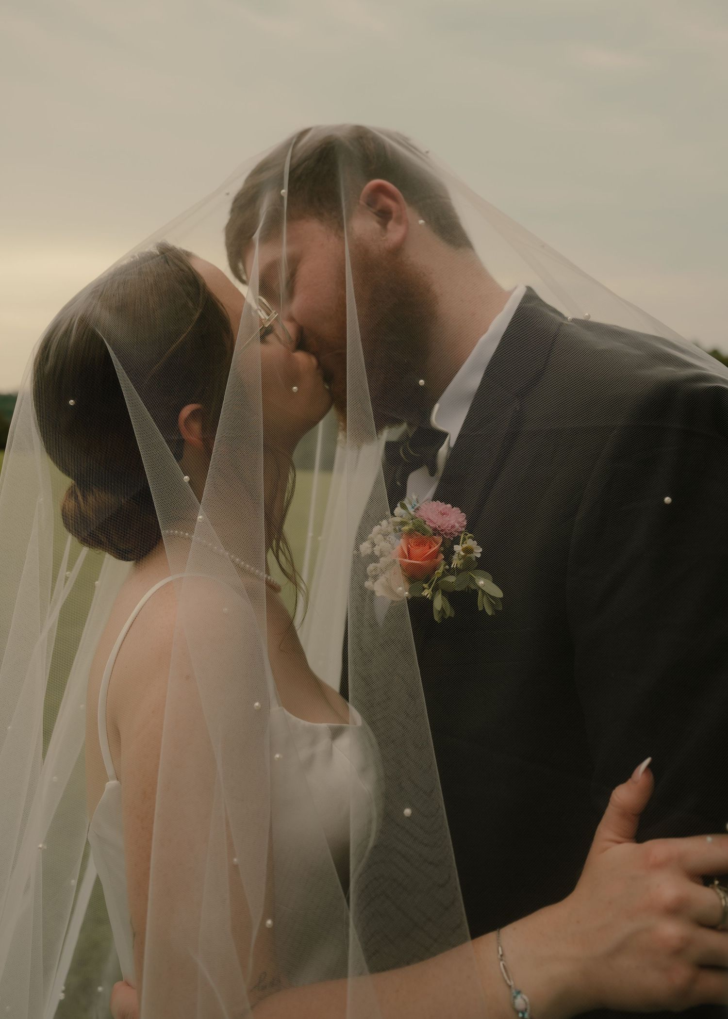 Bride and groom kissing under pearl-adorned veil with floral boutonniere during outdoor wedding.