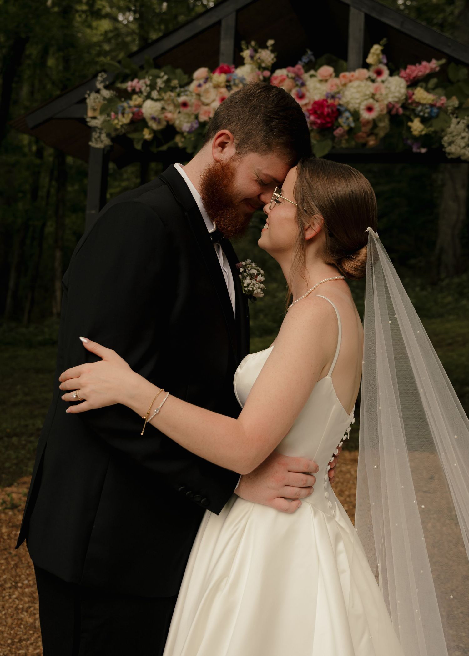 Bride and groom share intimate forehead touch moment before colorful floral arch in wooded ceremony setting.