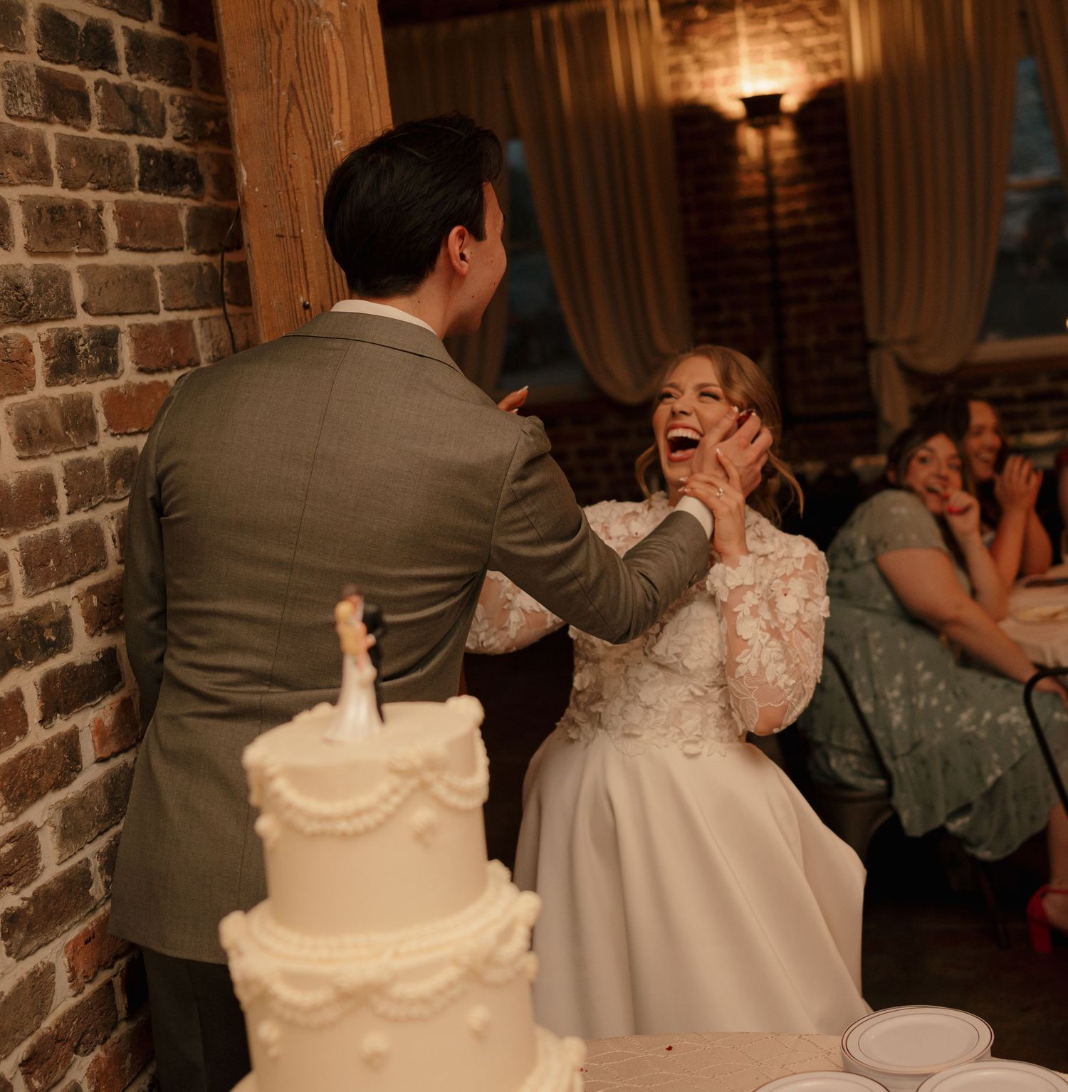 Groom playfully smearing wedding cake on laughing bride's face at rustic reception venue.