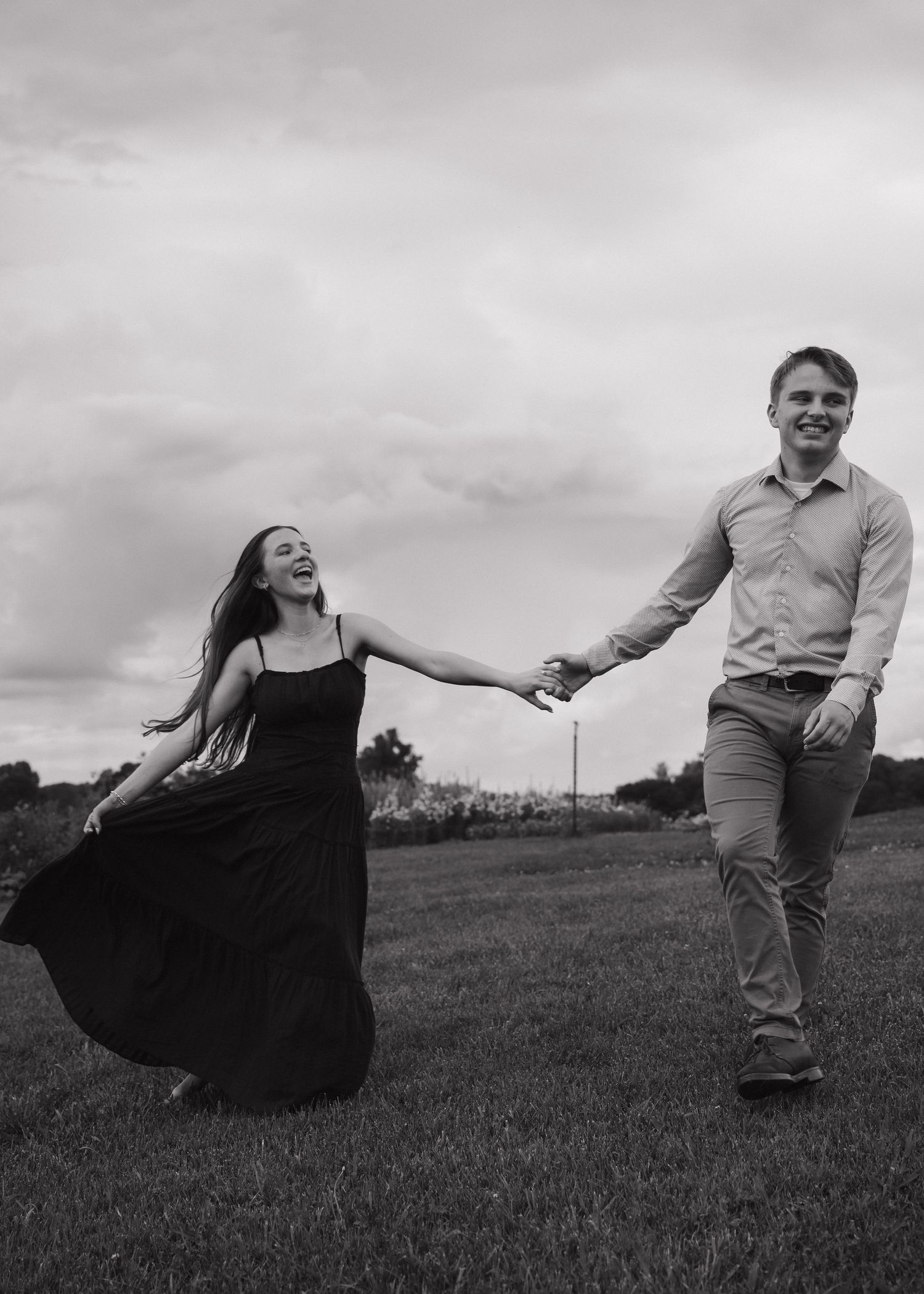 Black and white photo of a joyful couple holding hands and running through a grassy field under a cloudy sky.