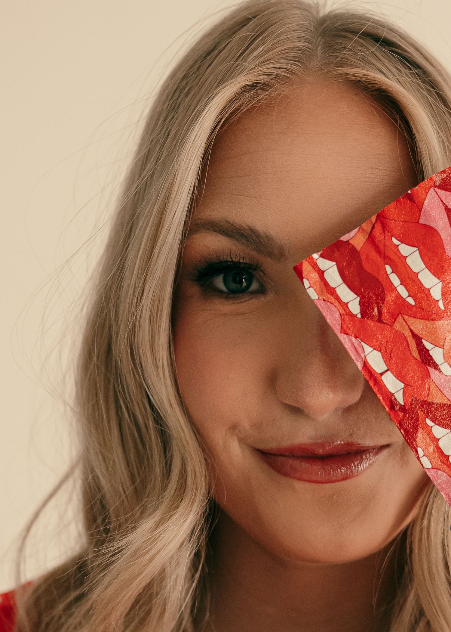 Blonde woman smiling, holding a red floral fabric over one eye, close-up portrait shot.