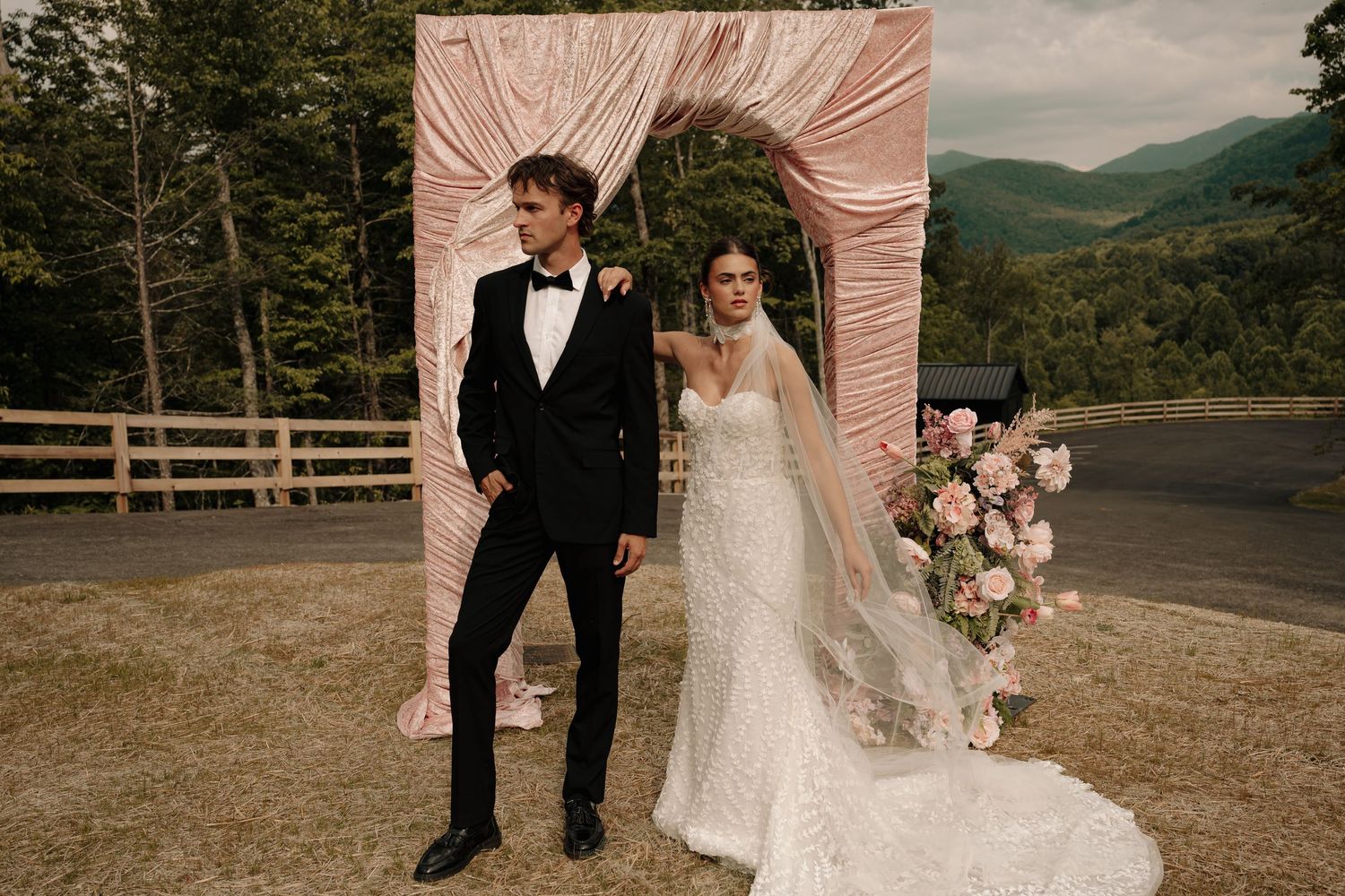 Bride and groom stand before a pink draped arch with mountain views and floral arrangements at an outdoor wedding ceremony.