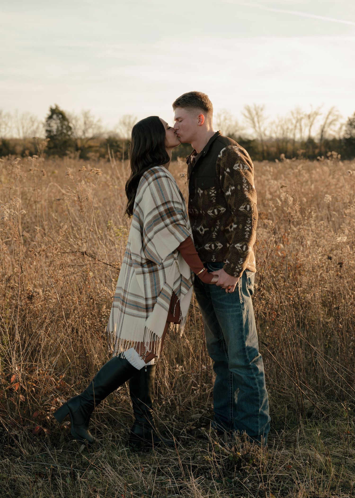 Couple kissing in golden field at sunset, woman in plaid poncho and knee-high boots holding hands with man in jeans.