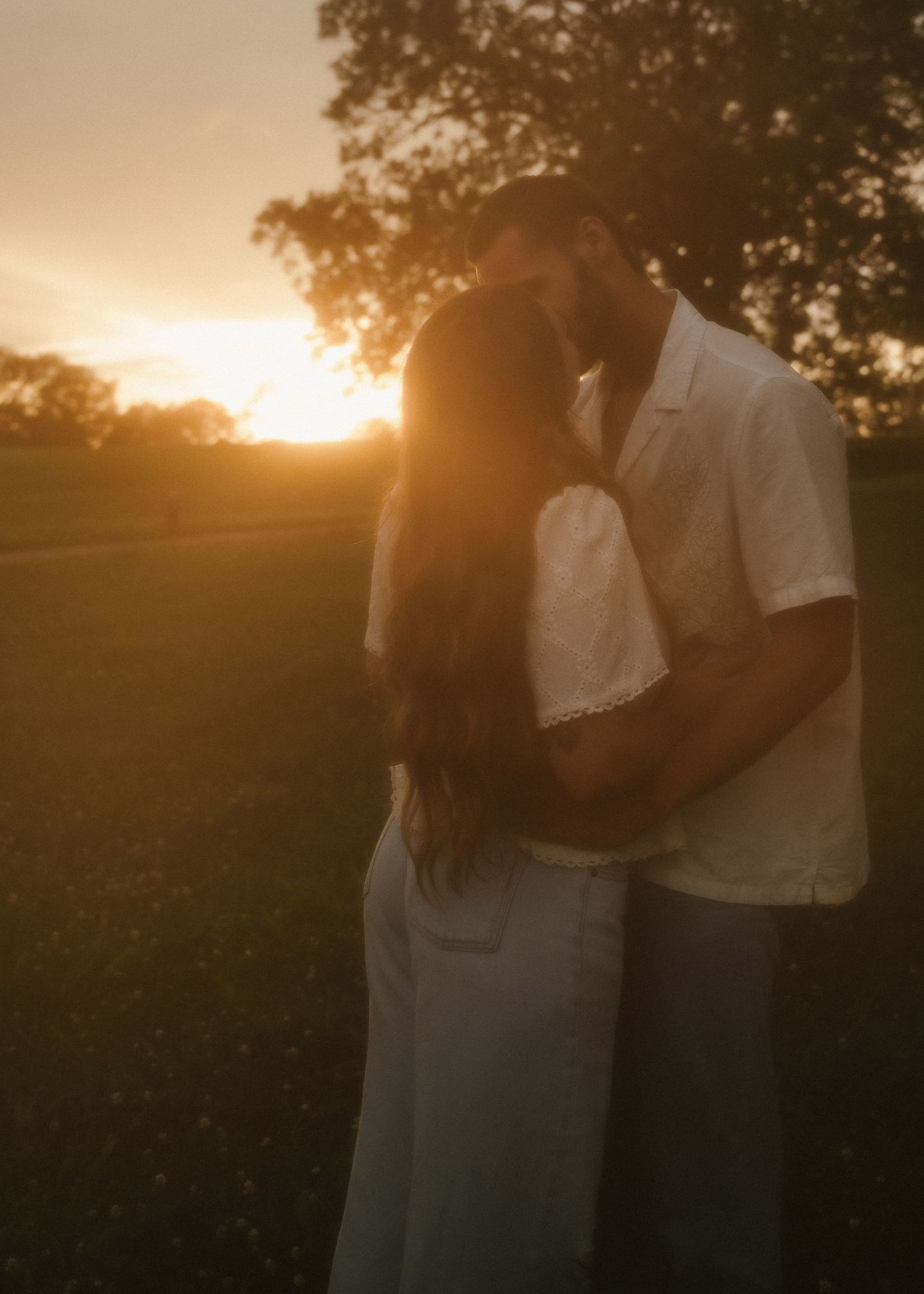 Romantic couple kissing at golden sunset with warm glowing light behind them outdoors.