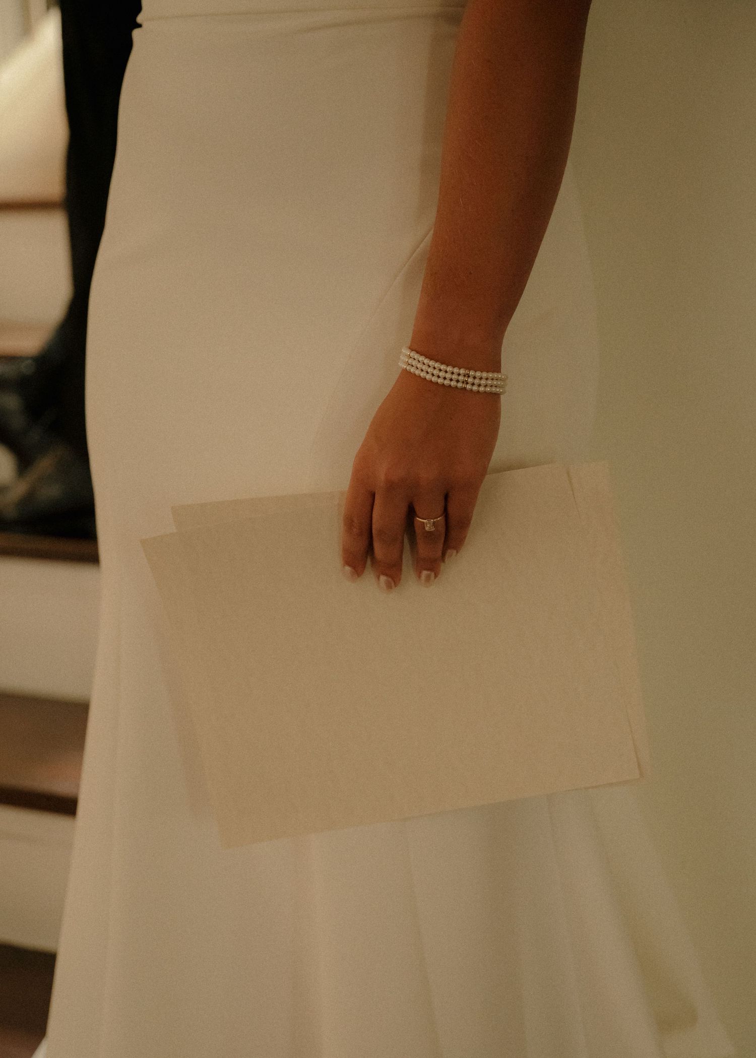 Close-up of bride holding paper in white wedding gown with pearl bracelet and diamond engagement ring.