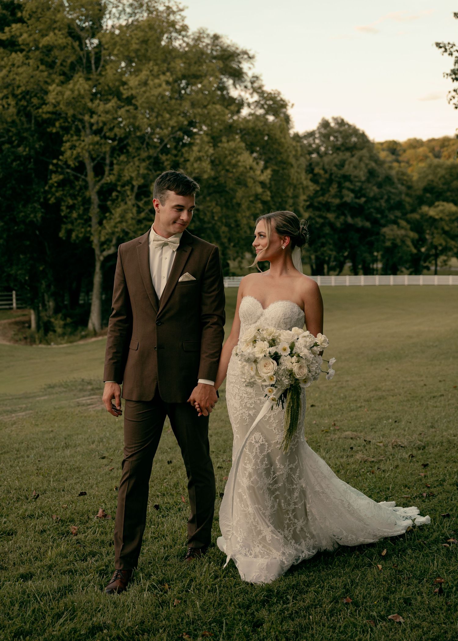 Bride and groom in brown suit hold hands on green lawn at sunset during elegant outdoor wedding portrait. Passerine Hills Estate Wedding TN