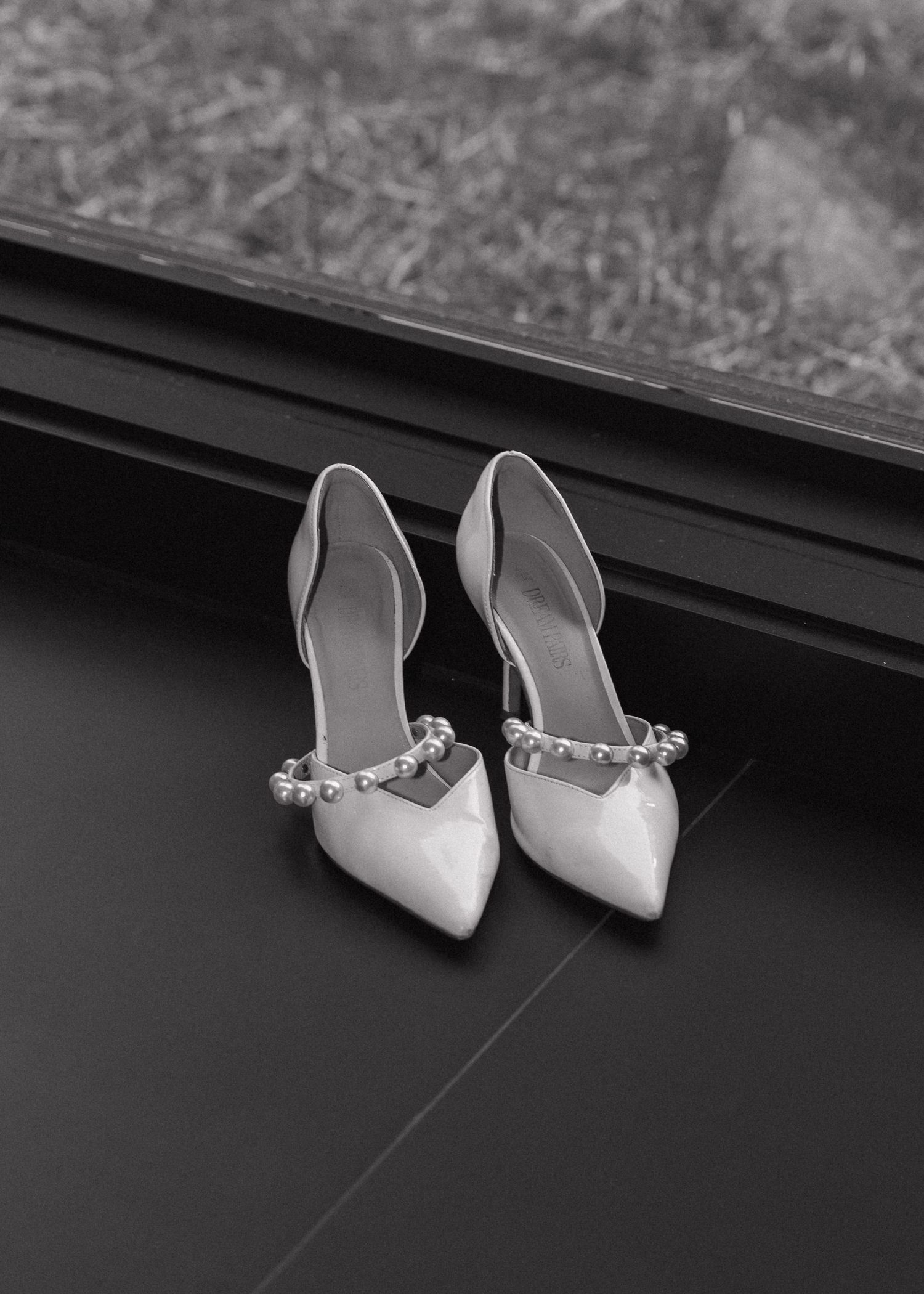 Black and white photo of elegant pointed-toe heels with pearl embellishments on a dark windowsill.