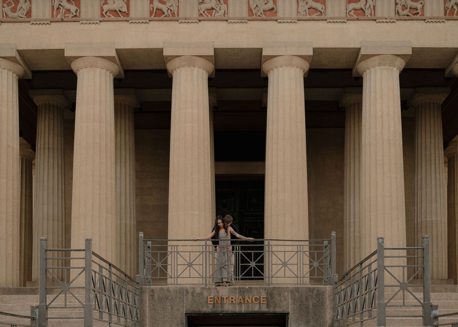 A couple stands on a balcony of the Nashville Parthenon, surrounded by towering Doric columns at dusk.