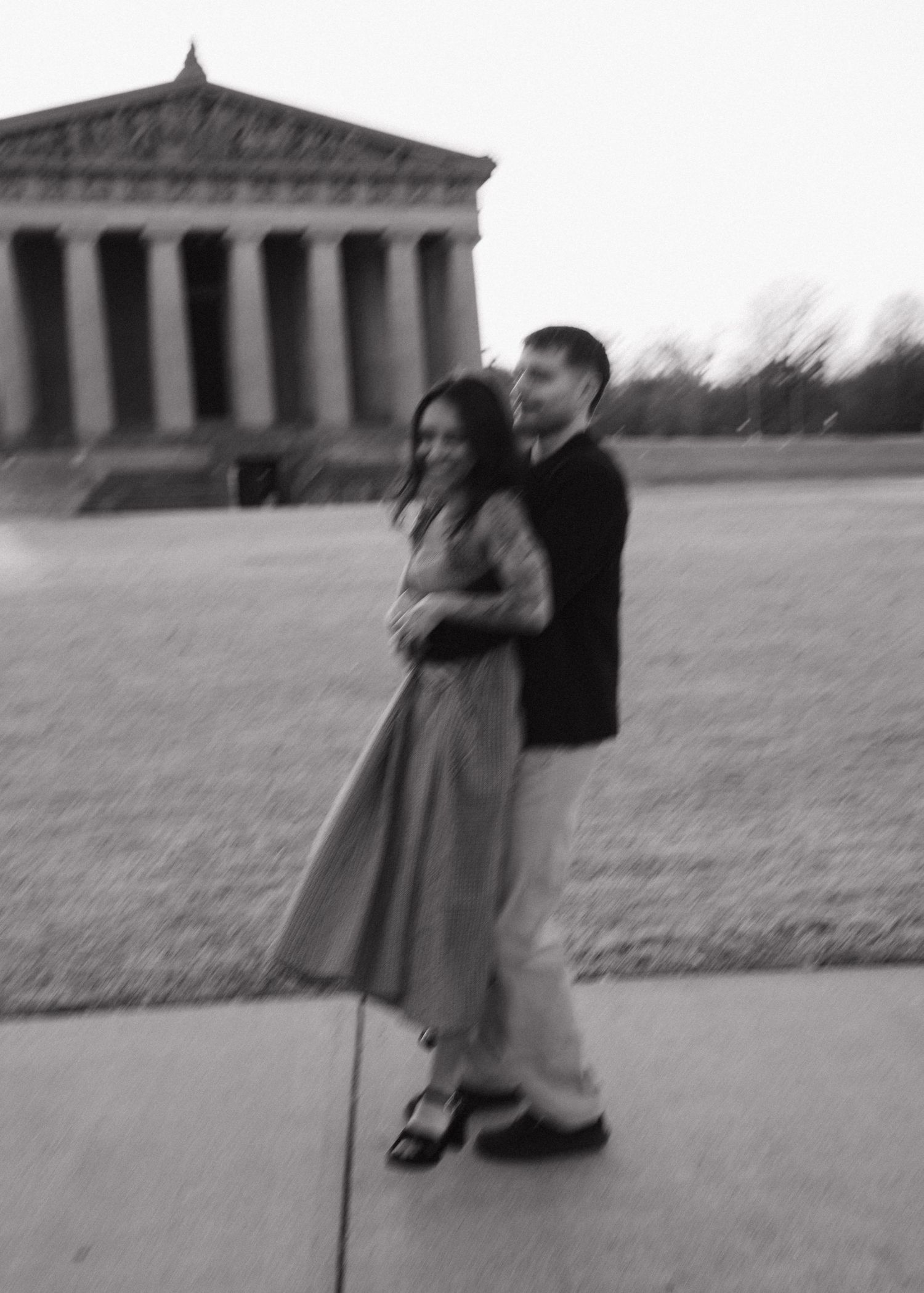 Blurry black and white photo of smiling couple embracing outside Greek temple.