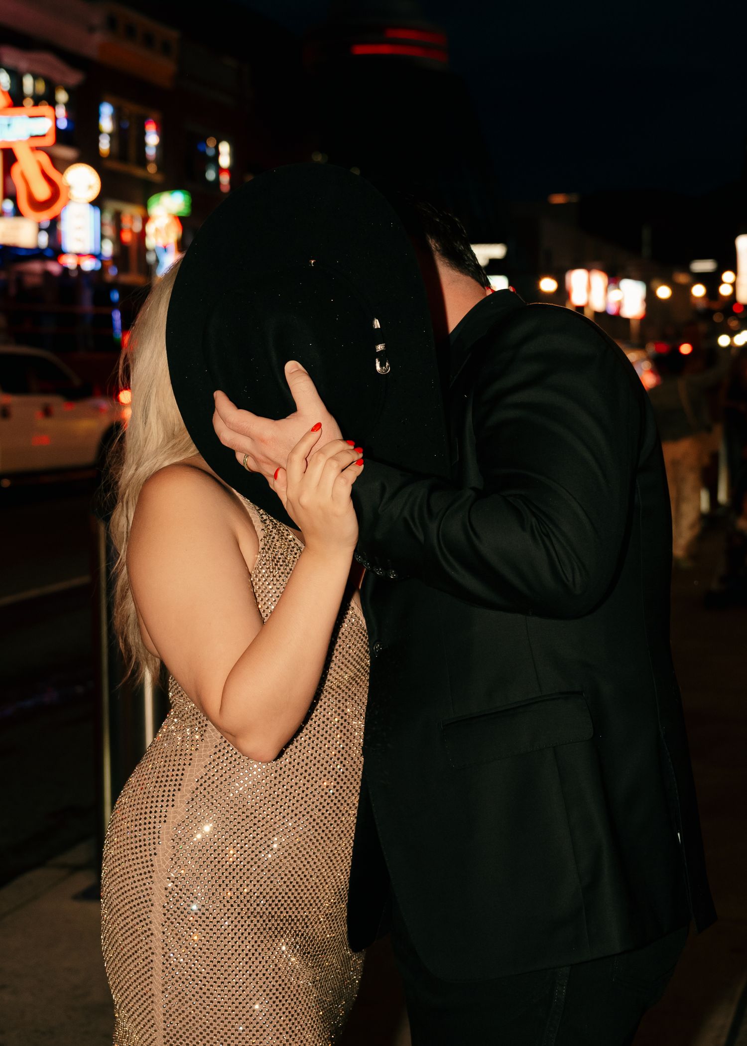 Couple kissing behind a black cowboy hat at night on a glittering city street, woman in sequin dress.