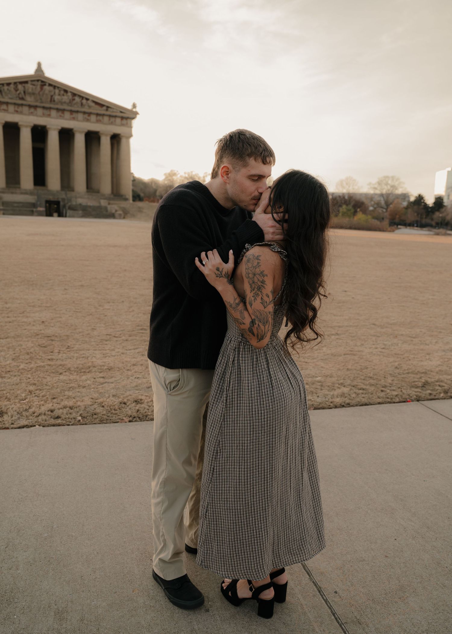 A couple shares a kiss in front of the Nashville Parthenon replica at dusk, the woman wearing a gingham dress with tattoos.