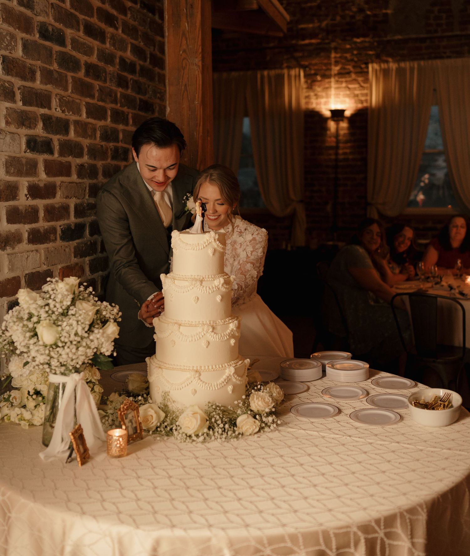Bride and groom cutting a tall white four-tier wedding cake at a rustic brick-walled reception.