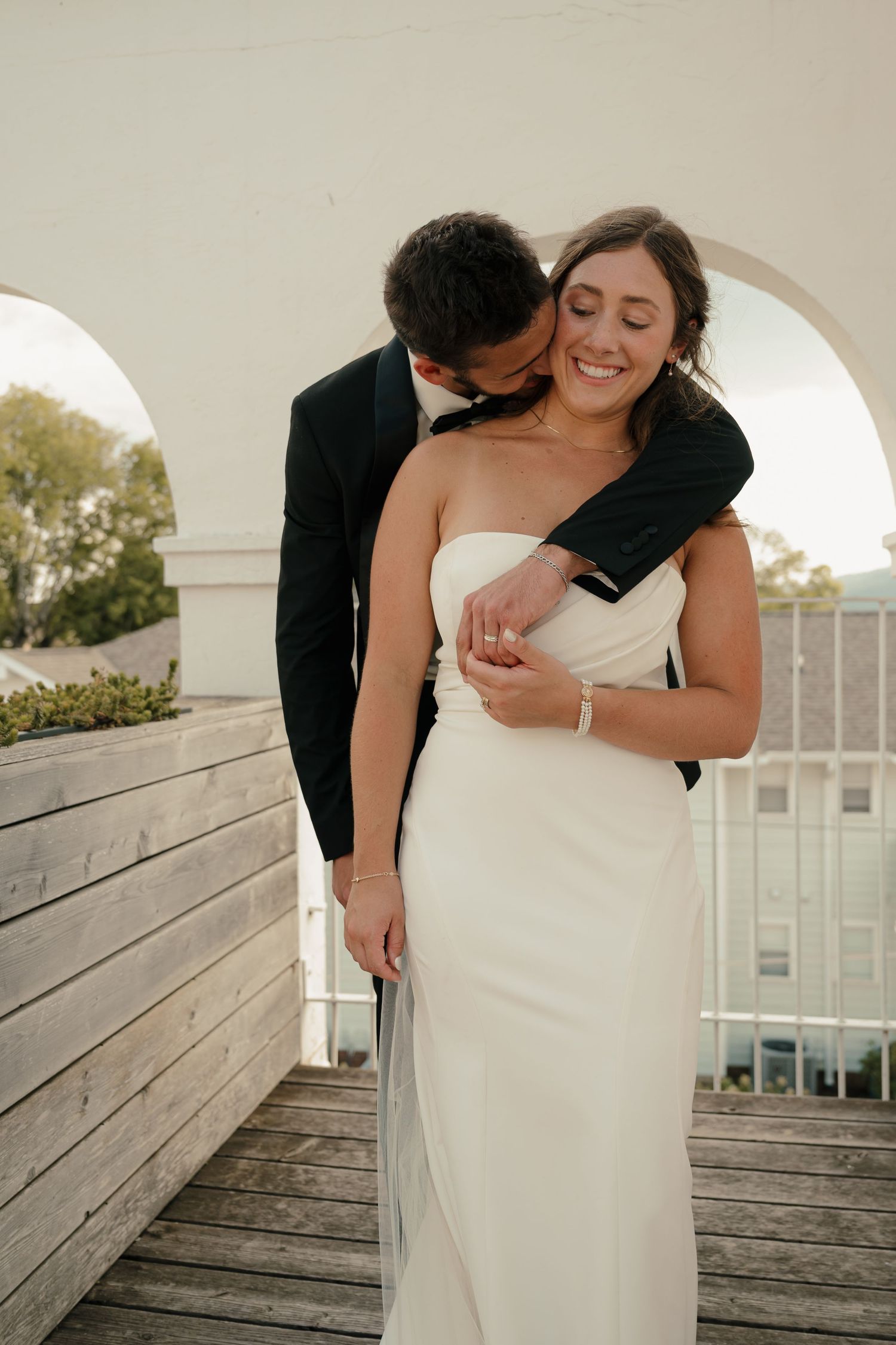 Groom in black suit kissing bride's neck as she smiles on rooftop balcony with white arches.