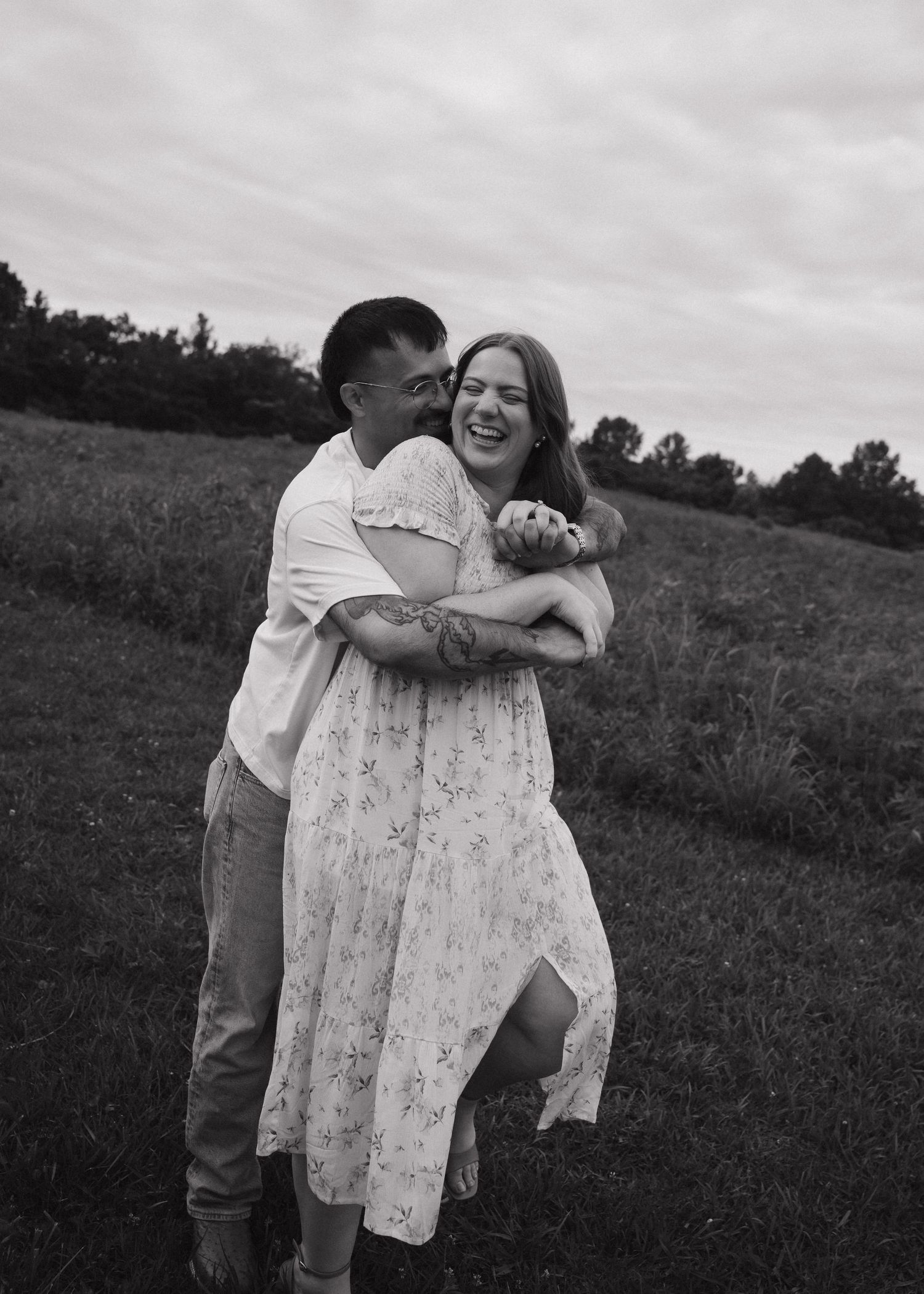 A joyful couple embraces and laughs together in a black and white photo taken in a grassy field.