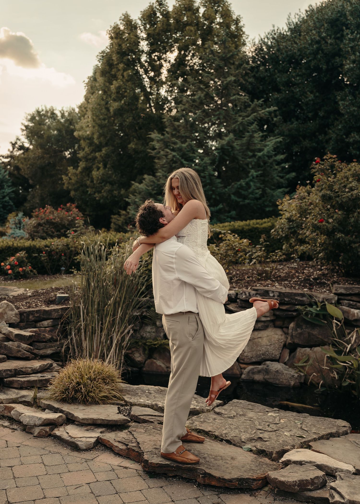 A man lifts a woman in a white dress in a lush garden setting during golden hour.