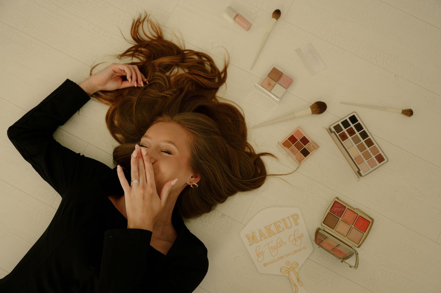 Woman with long red hair lying on white floor surrounded by makeup products and beauty accessories.