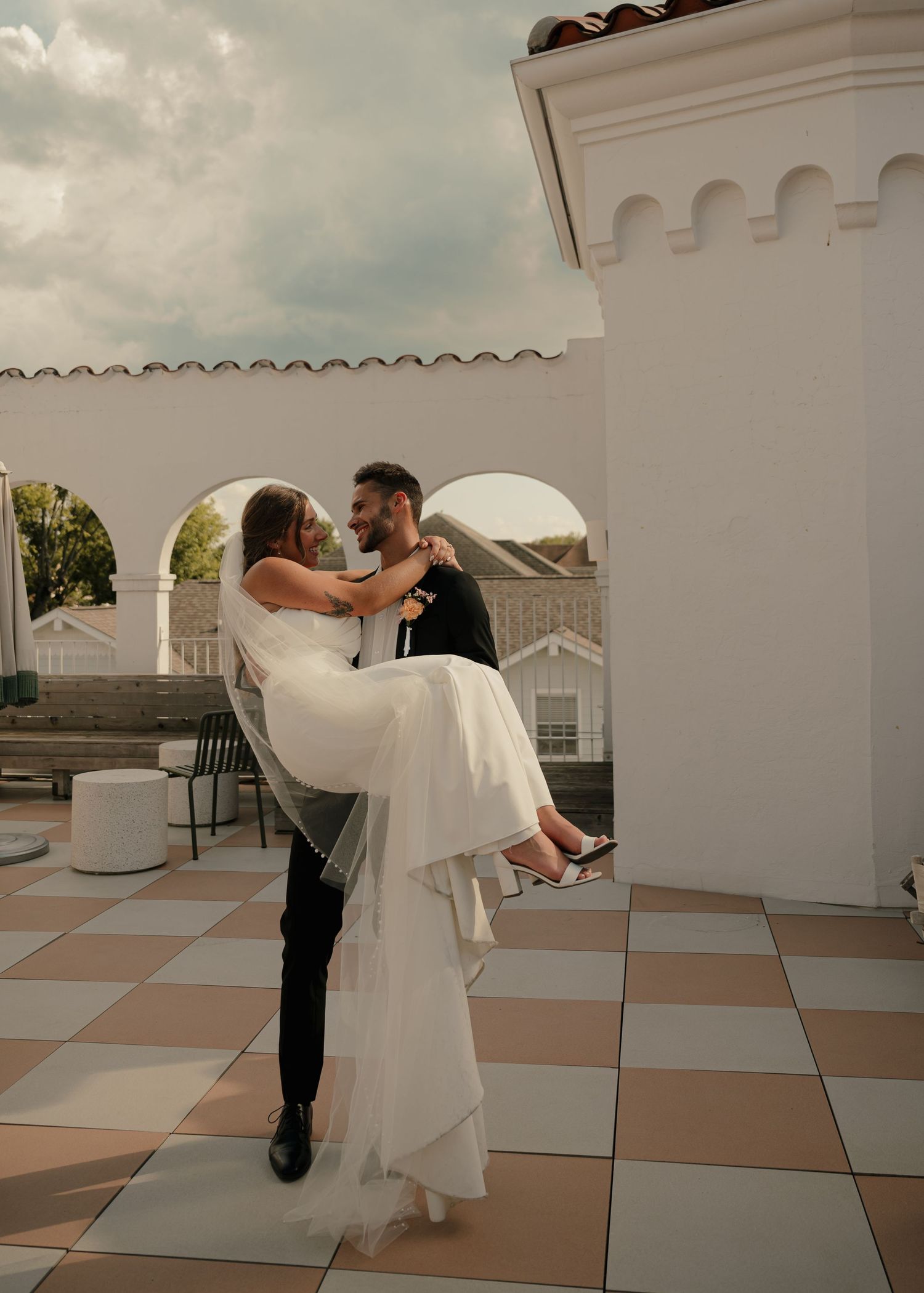 Groom lifting bride in white wedding gown on checkered rooftop terrace with white arches.