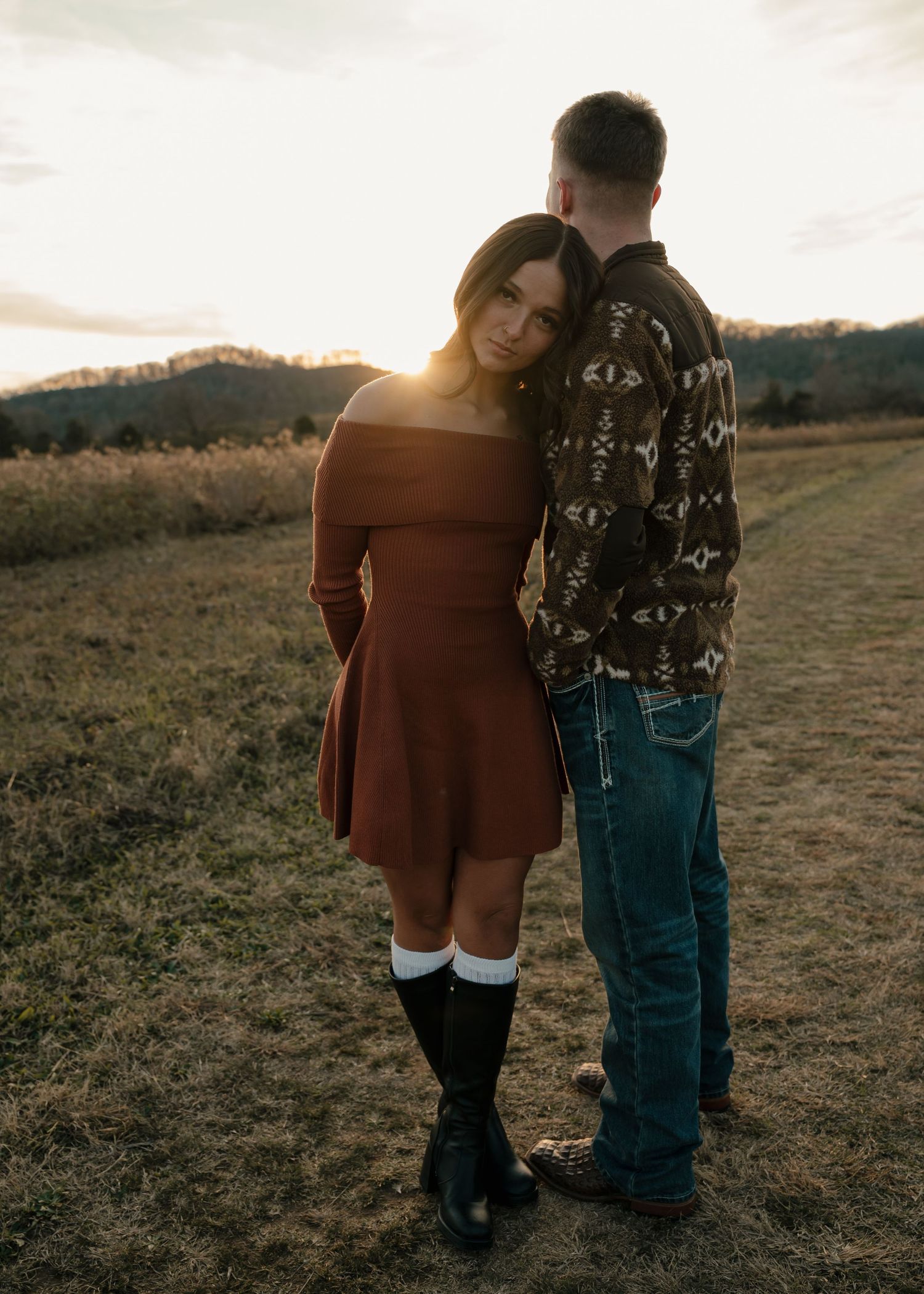Woman in rust off-shoulder dress leaning on mans shoulder at golden hour in an open countryside field.