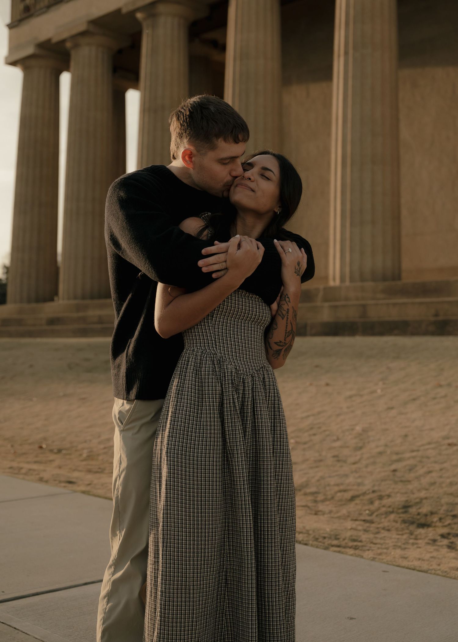 Man kissing woman on cheek while embracing near ancient Greek-style columns.