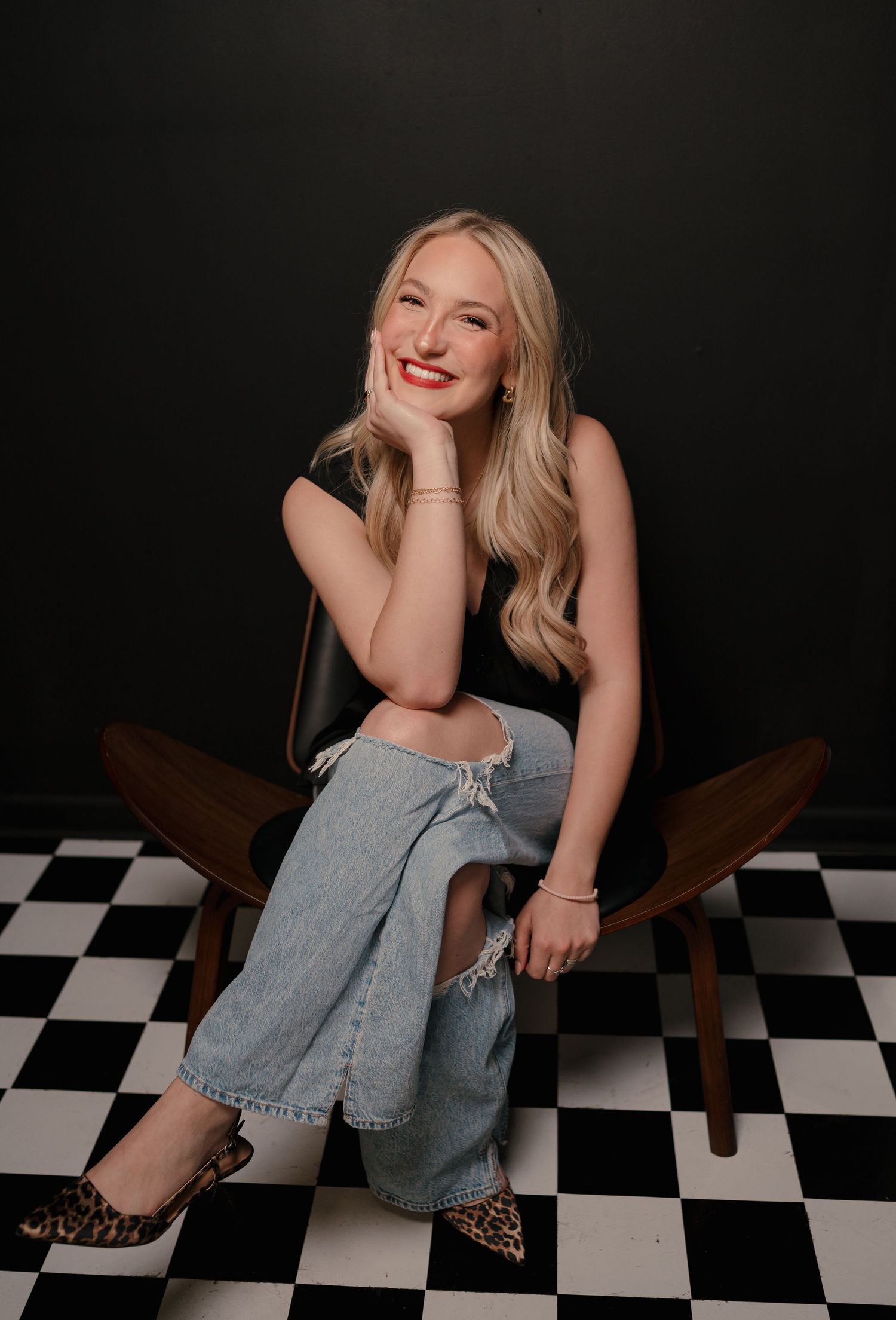 Blonde woman in black top and jeans sitting on chair with checkered floor.