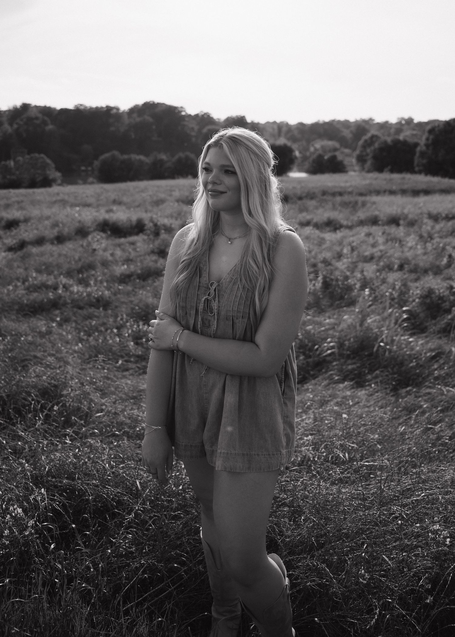 Young blonde woman smiling in a black and white photo, standing in a sunlit field wearing a fringe dress and boots.