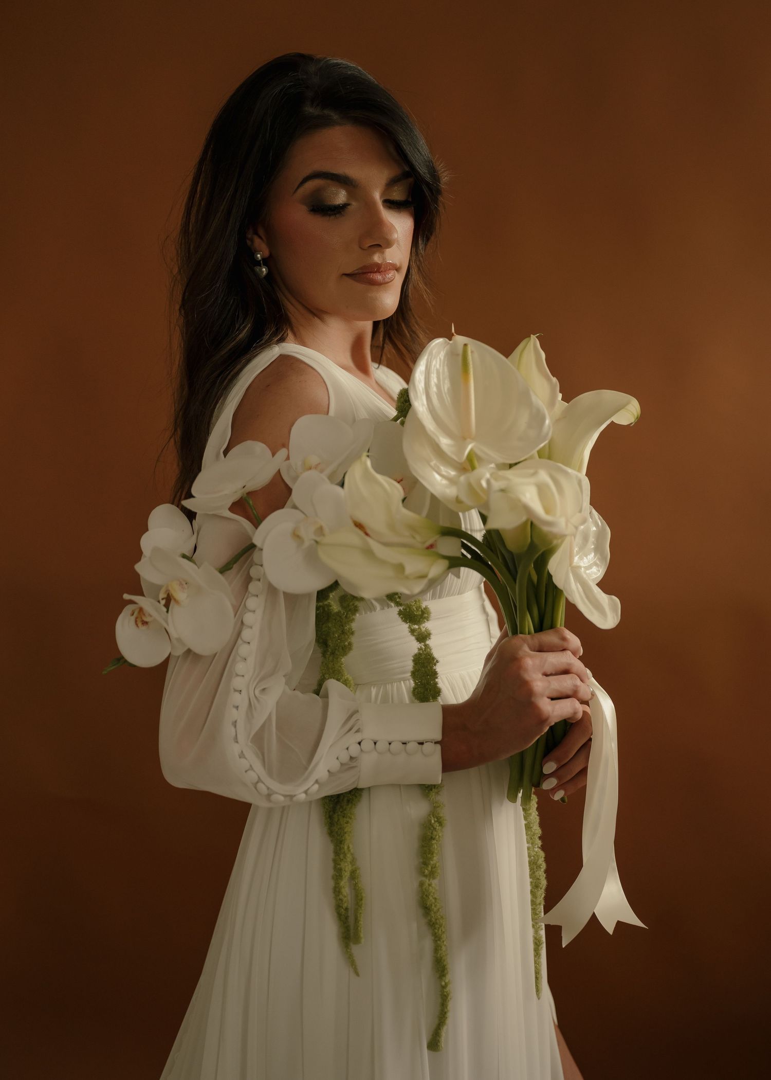 Elegant woman in white dress holding bouquet of white calla lilies and orchids against brown background.