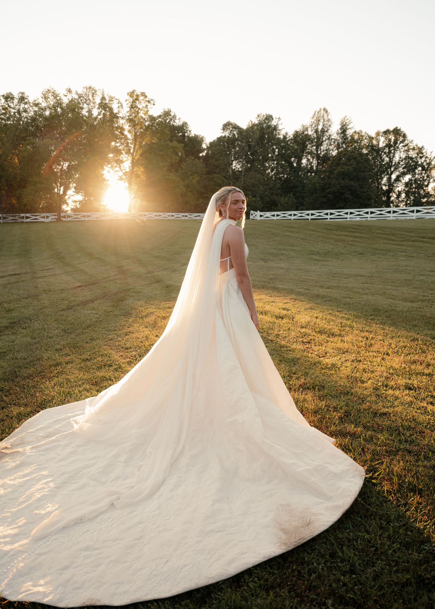 Bride in flowing white gown with long veil standing in sunlit green field at golden hour.