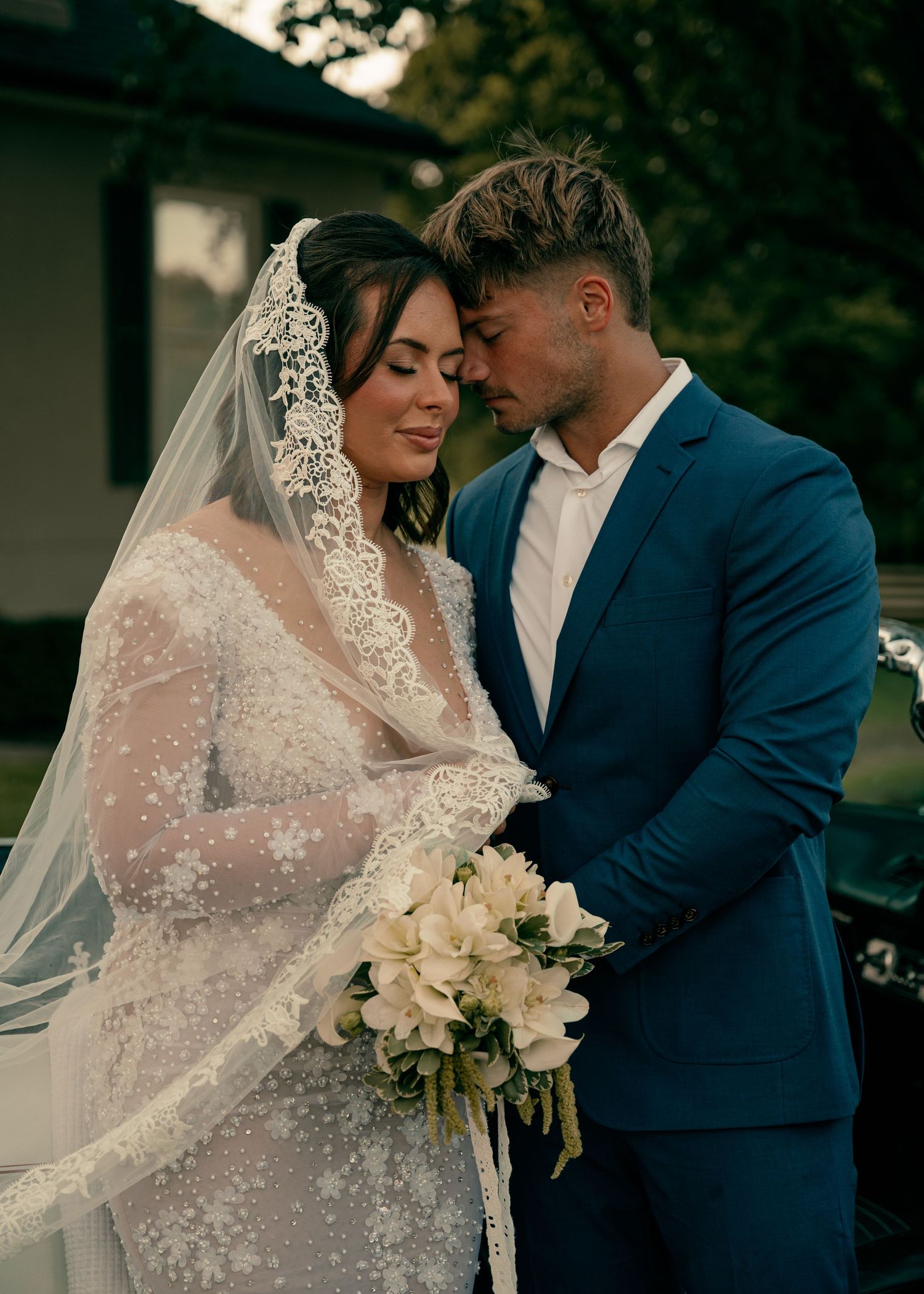 Bride in lace wedding dress with veil and groom in blue suit embracing outdoors with white bouquet.