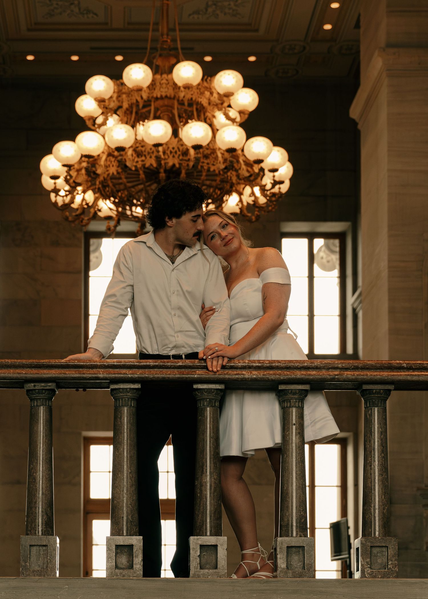Couple embracing on elegant balcony beneath ornate chandelier, woman in white dress leaning on man in white shirt.