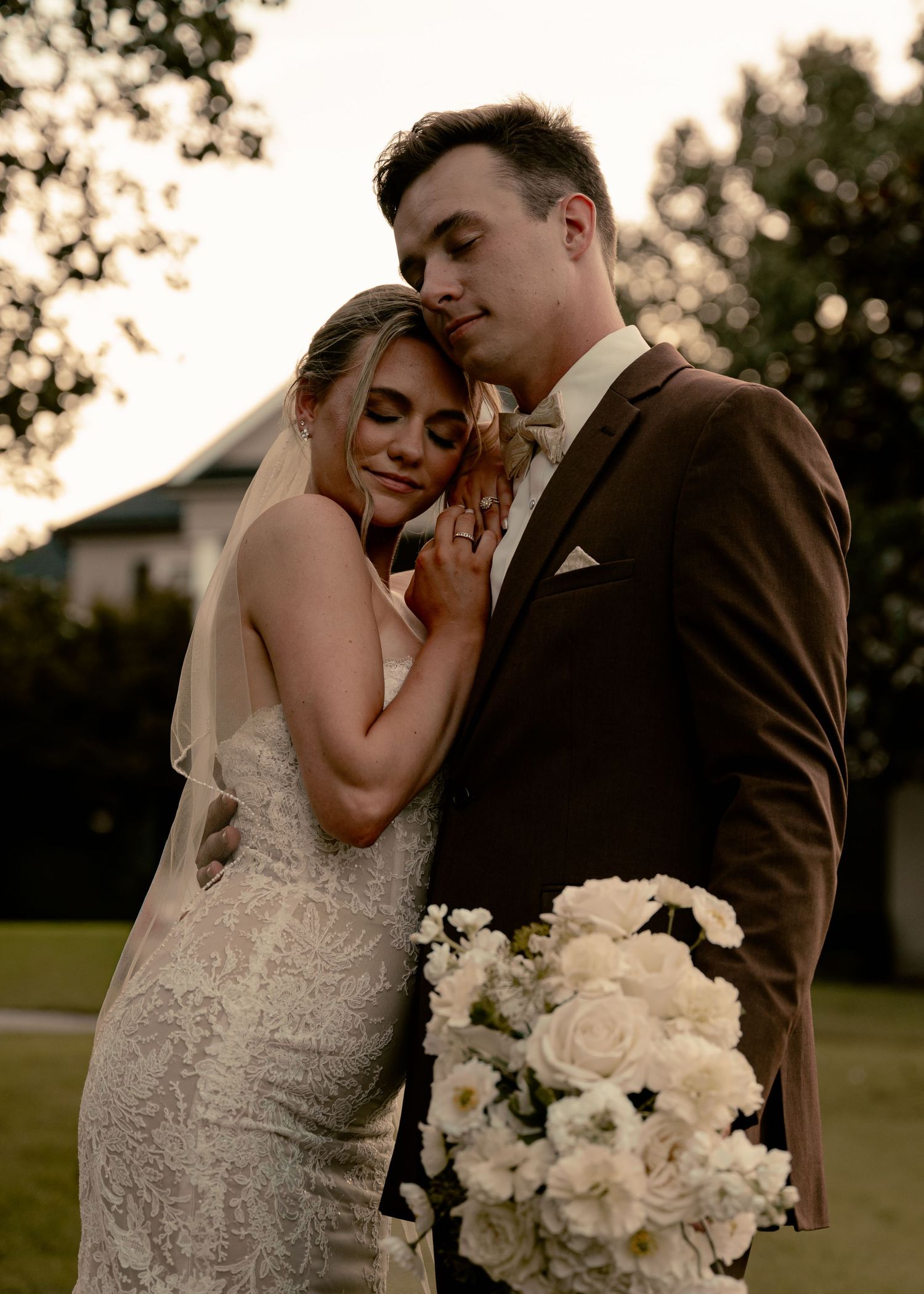 Bride in lace gown leans on groom in brown suit holding white floral bouquet at golden hour outdoor wedding. Passerine Hills Estate TN Wed