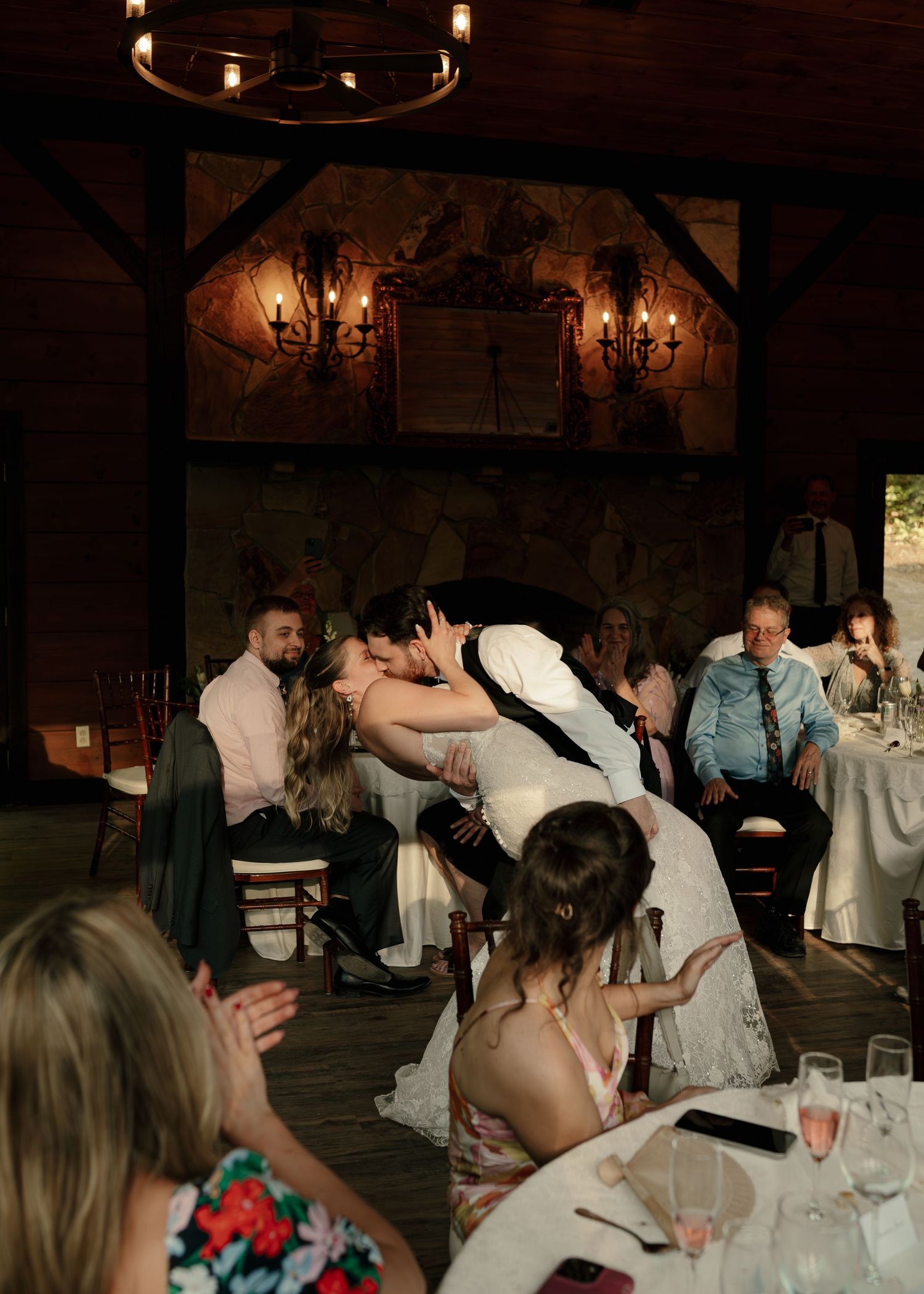 Bride and groom share romantic dip kiss at rustic wedding reception with stone fireplace.