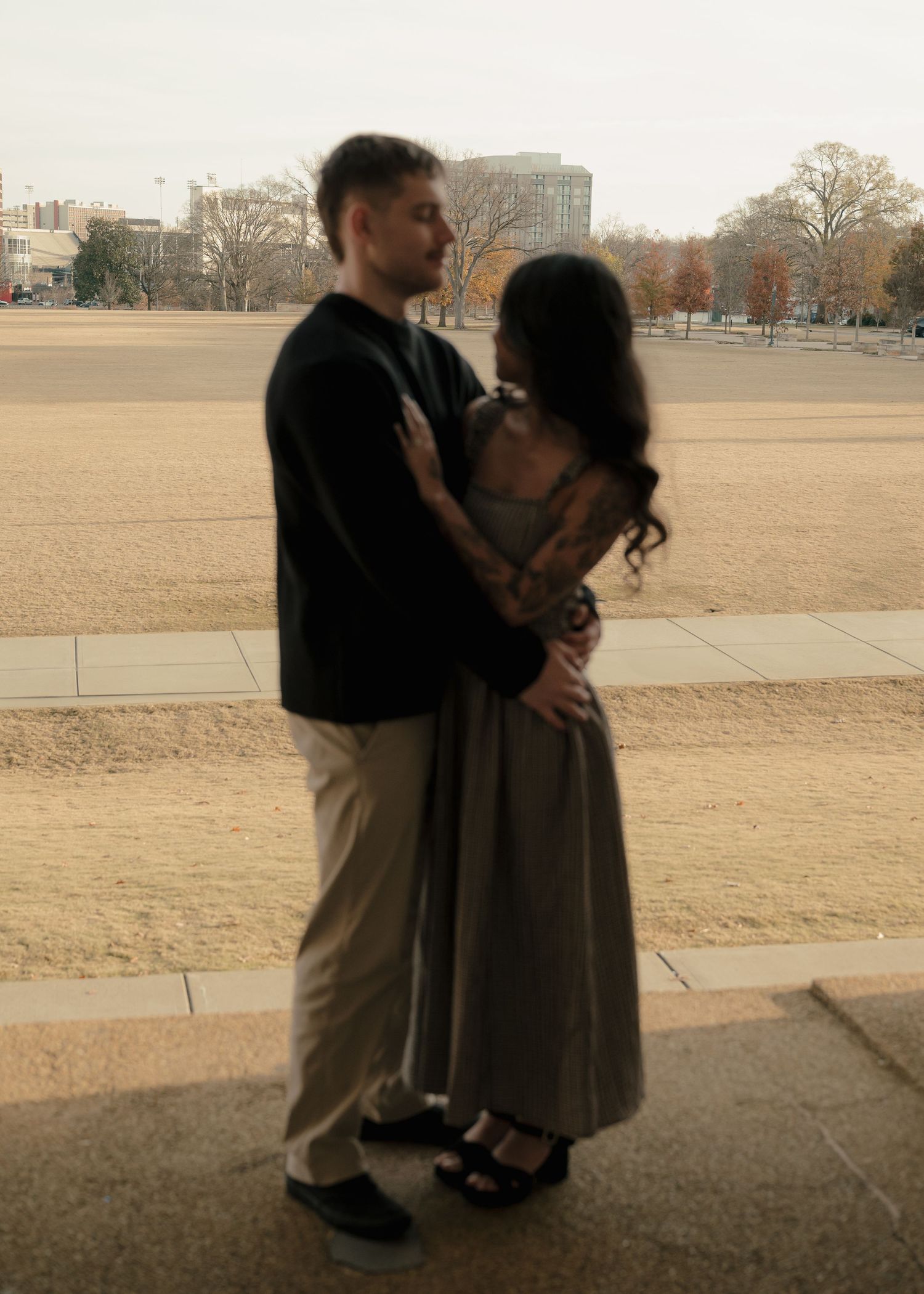 Couple embracing romantically outdoors near open field with city buildings in background.