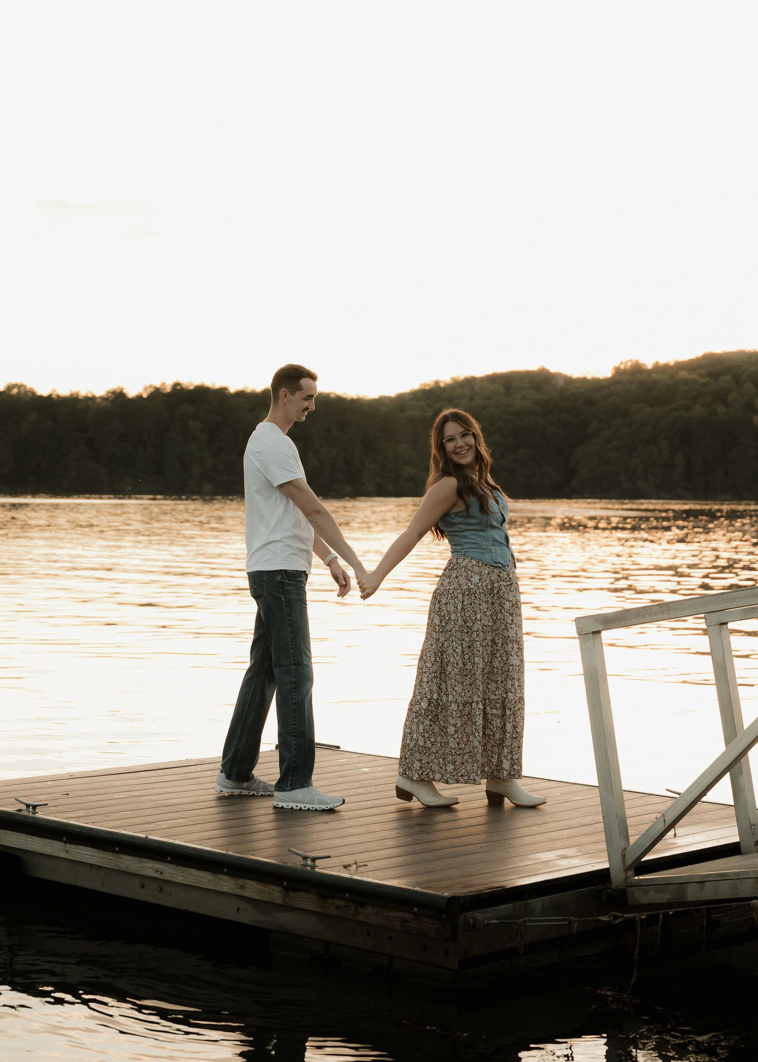 Happy couple holding hands and smiling on a wooden dock at golden sunset.