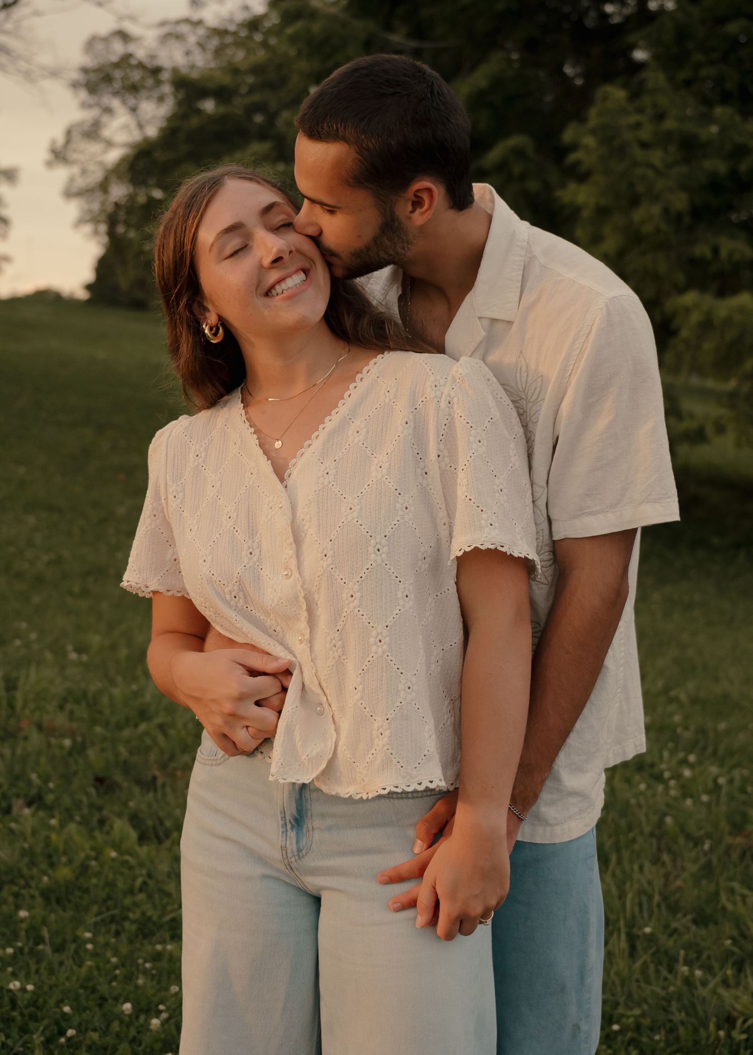 Man kissing woman on cheek as she laughs joyfully in a lush green outdoor setting.