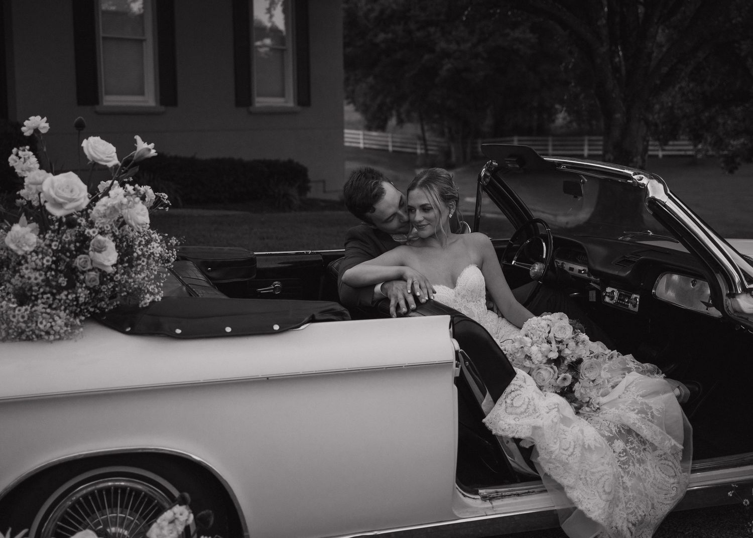 Black and white photo of bride and groom embracing in vintage convertible car decorated with flowers.