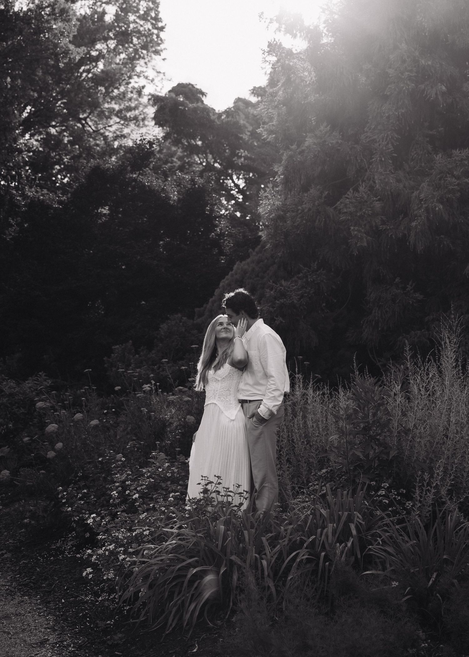 Black and white photo of a couple sharing a romantic moment surrounded by lush garden greenery and soft backlight.