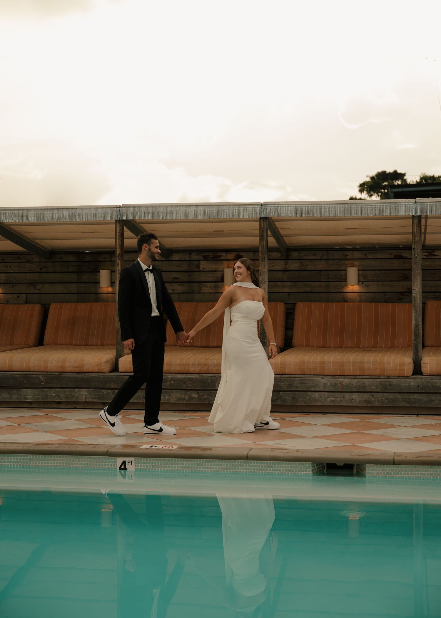 Bride and groom holding hands walking beside a pool at sunset during wedding reception.
