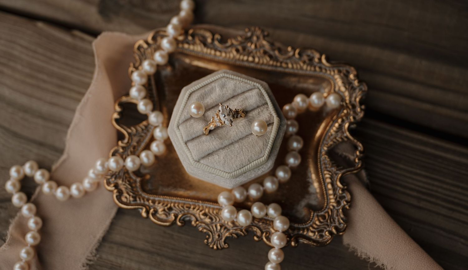 Elegant engagement ring displayed in a velvet box surrounded by pearl necklace on wood.