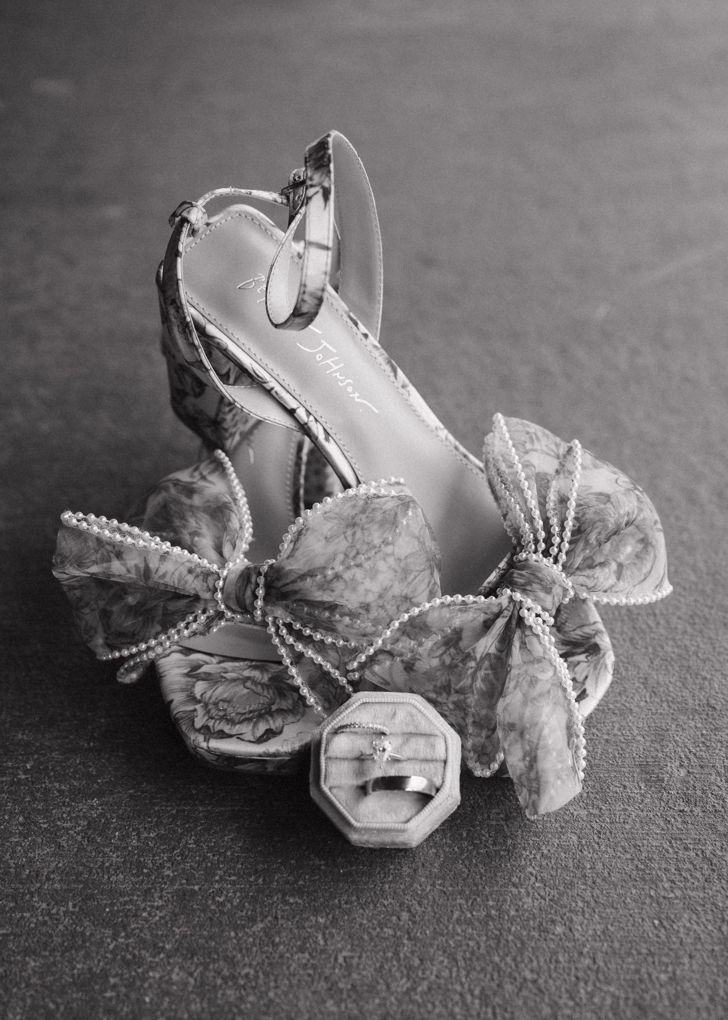 Black and white photo of elegant floral heels with pearl bow detail and wedding ring box on gray surface.
