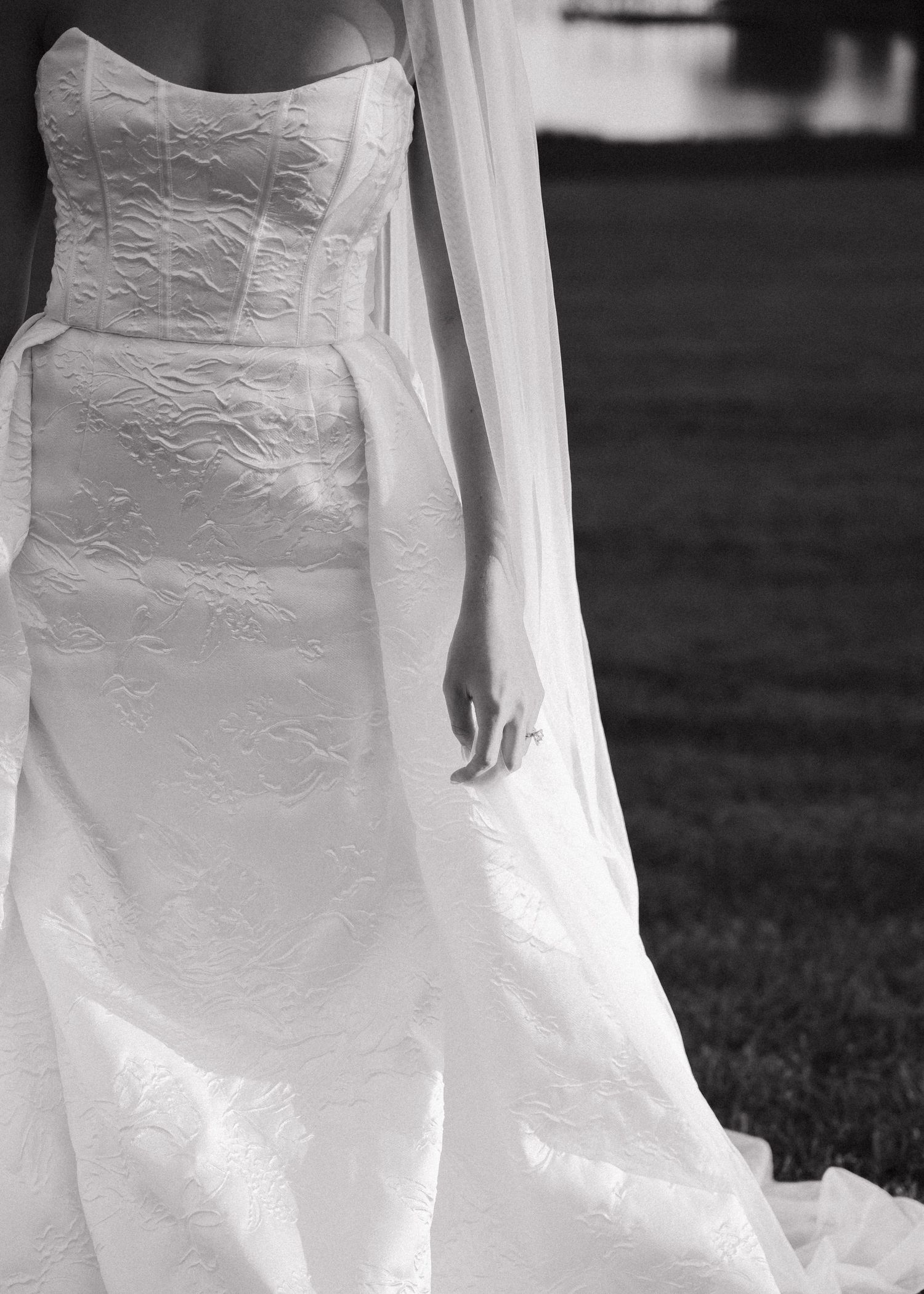 Black and white close-up of bride in strapless embossed wedding gown with flowing veil outdoors.