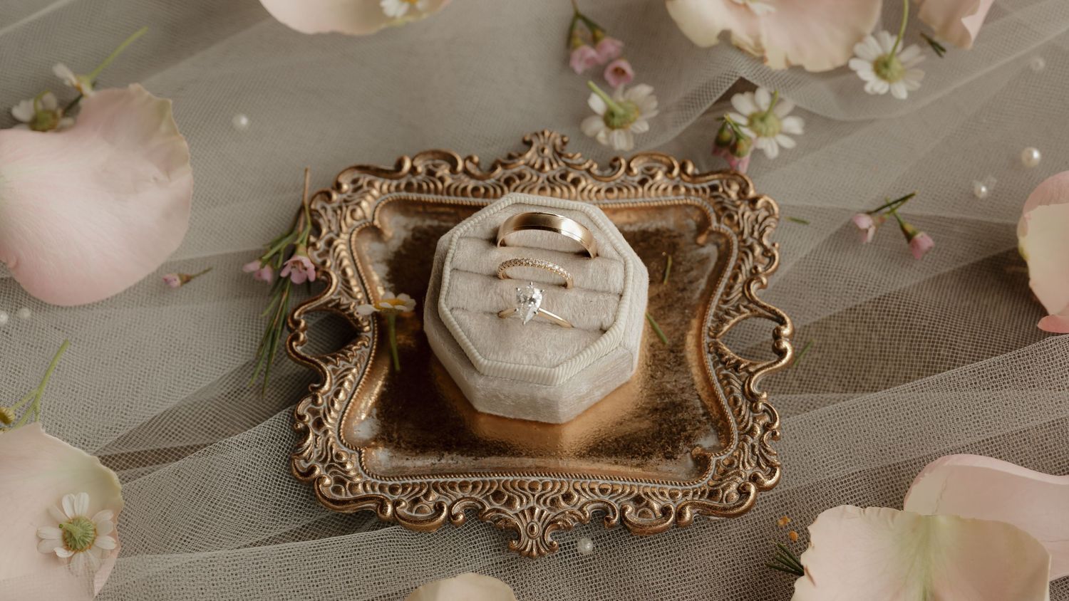 Gold wedding bands in velvet ring box on ornate gold frame surrounded by pink rose petals and florals.