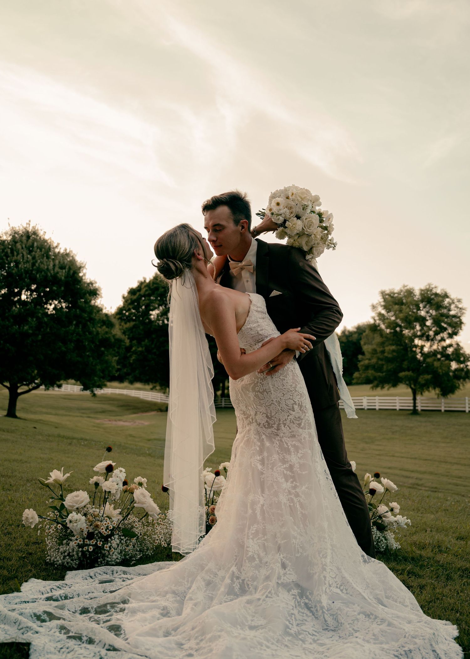 Bride and groom kissing at outdoor ceremony altar with white floral arrangements at golden hour.