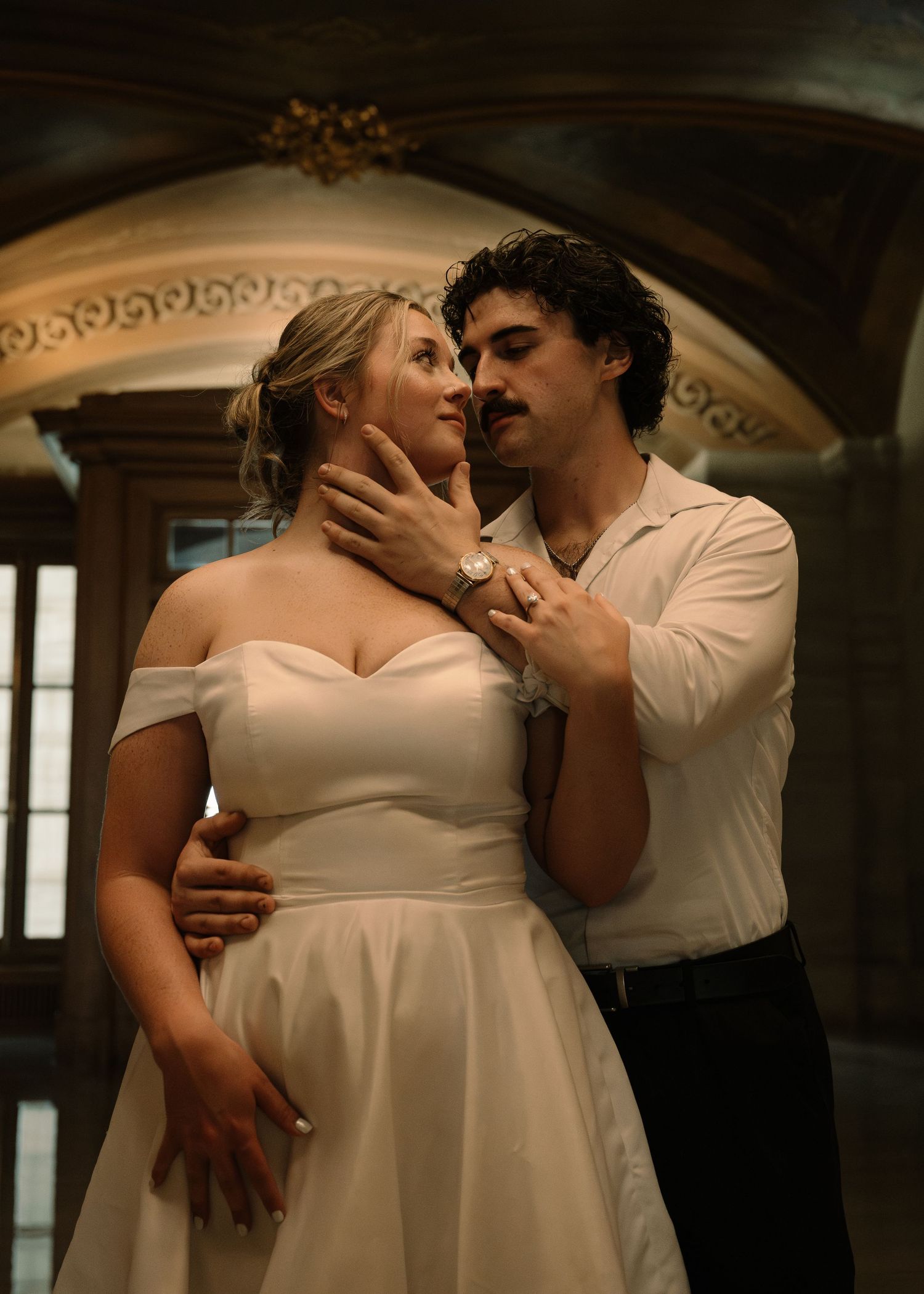 Romantic couple in wedding attire embrace tenderly in an elegant arched interior, bride in off-shoulder gown.