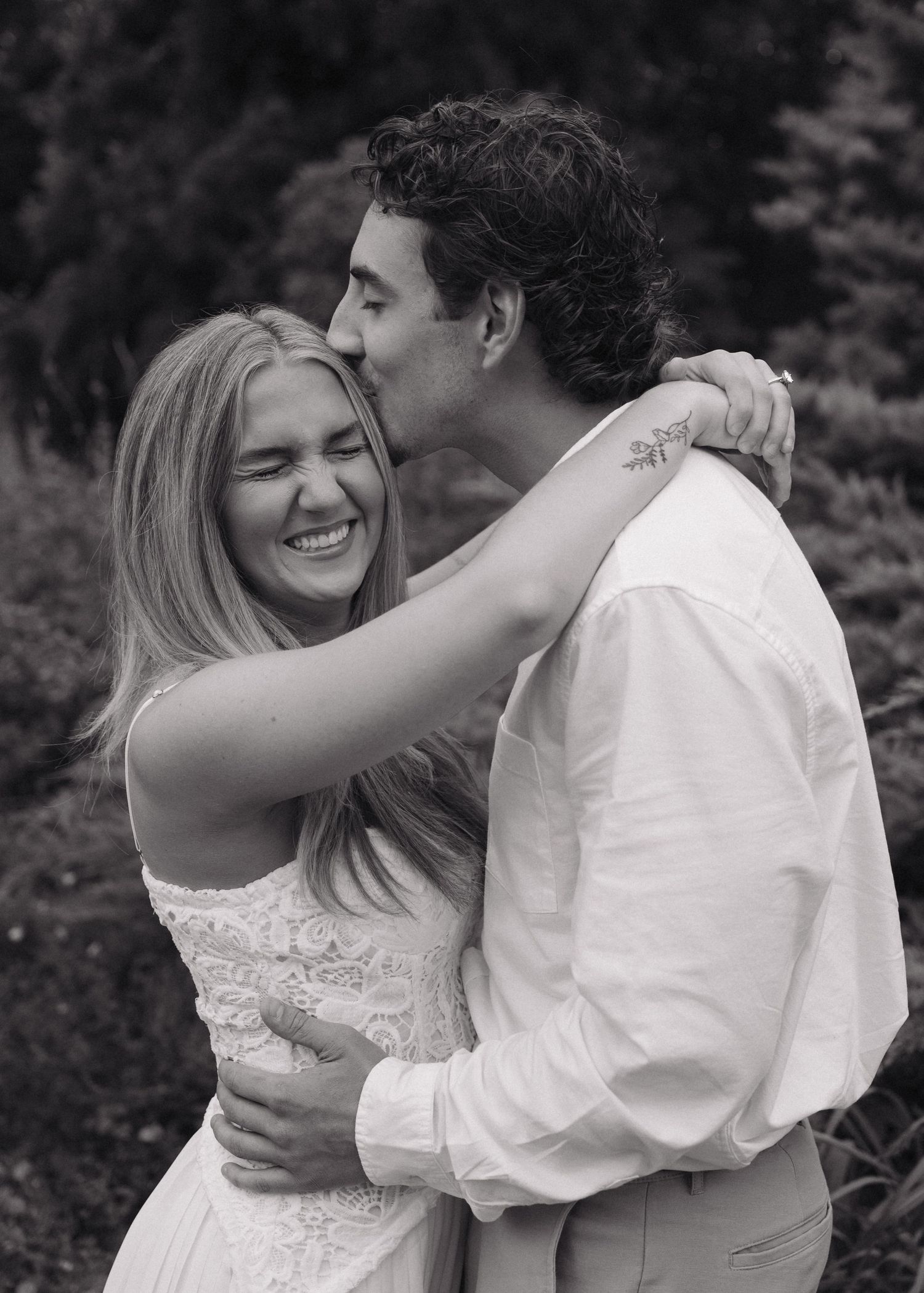 Black and white photo of a smiling bride and groom embracing outdoors, man kissing woman on cheek.