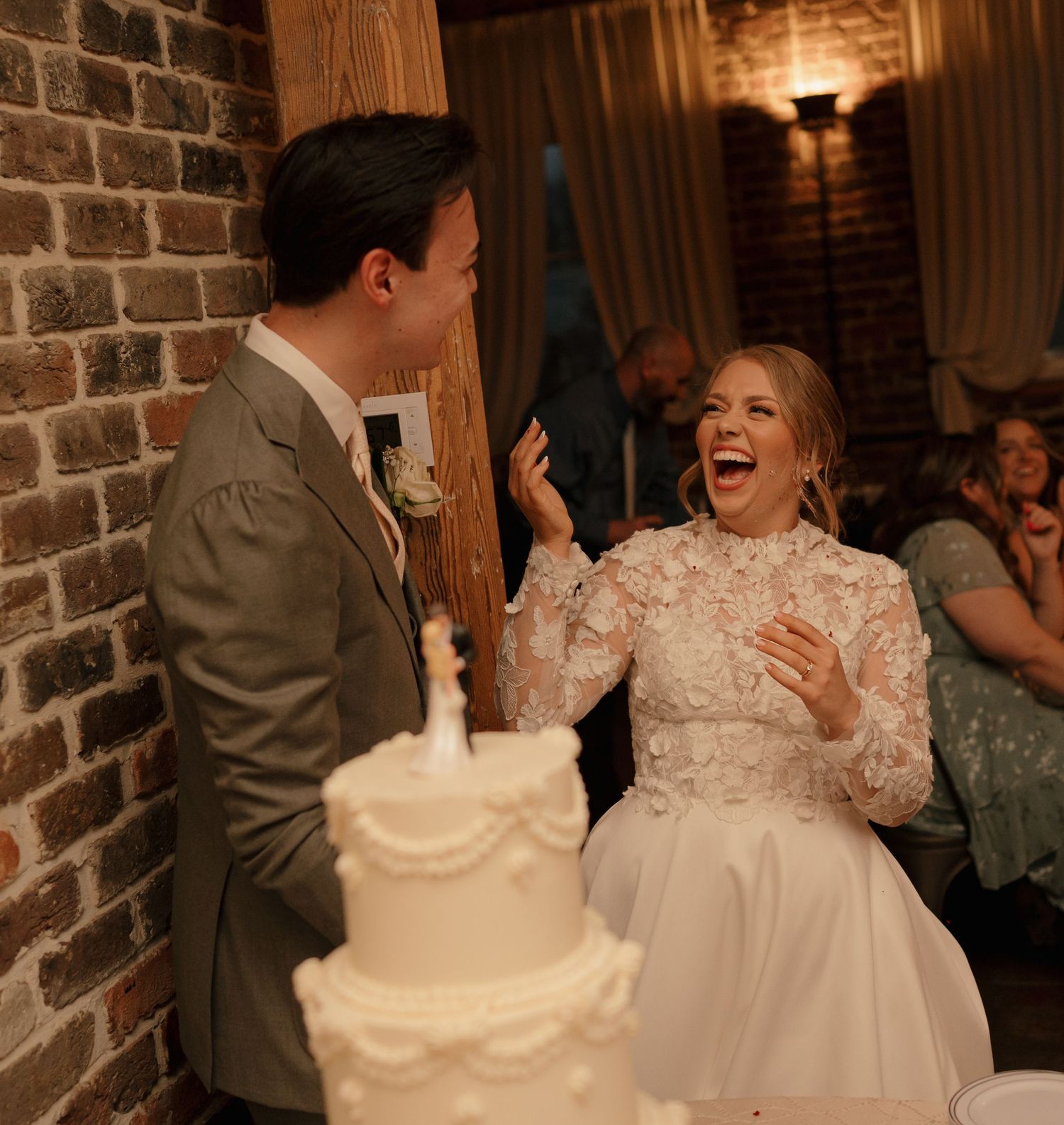 Bride and groom laughing together beside a white wedding cake in a rustic brick venue.