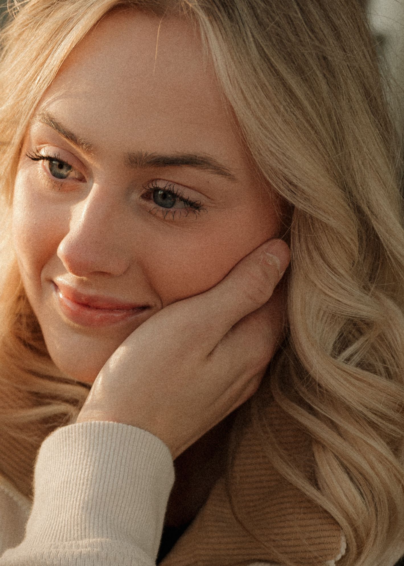 A close-up portrait of a smiling blonde woman with blue eyes resting her cheek gently on her hand.
