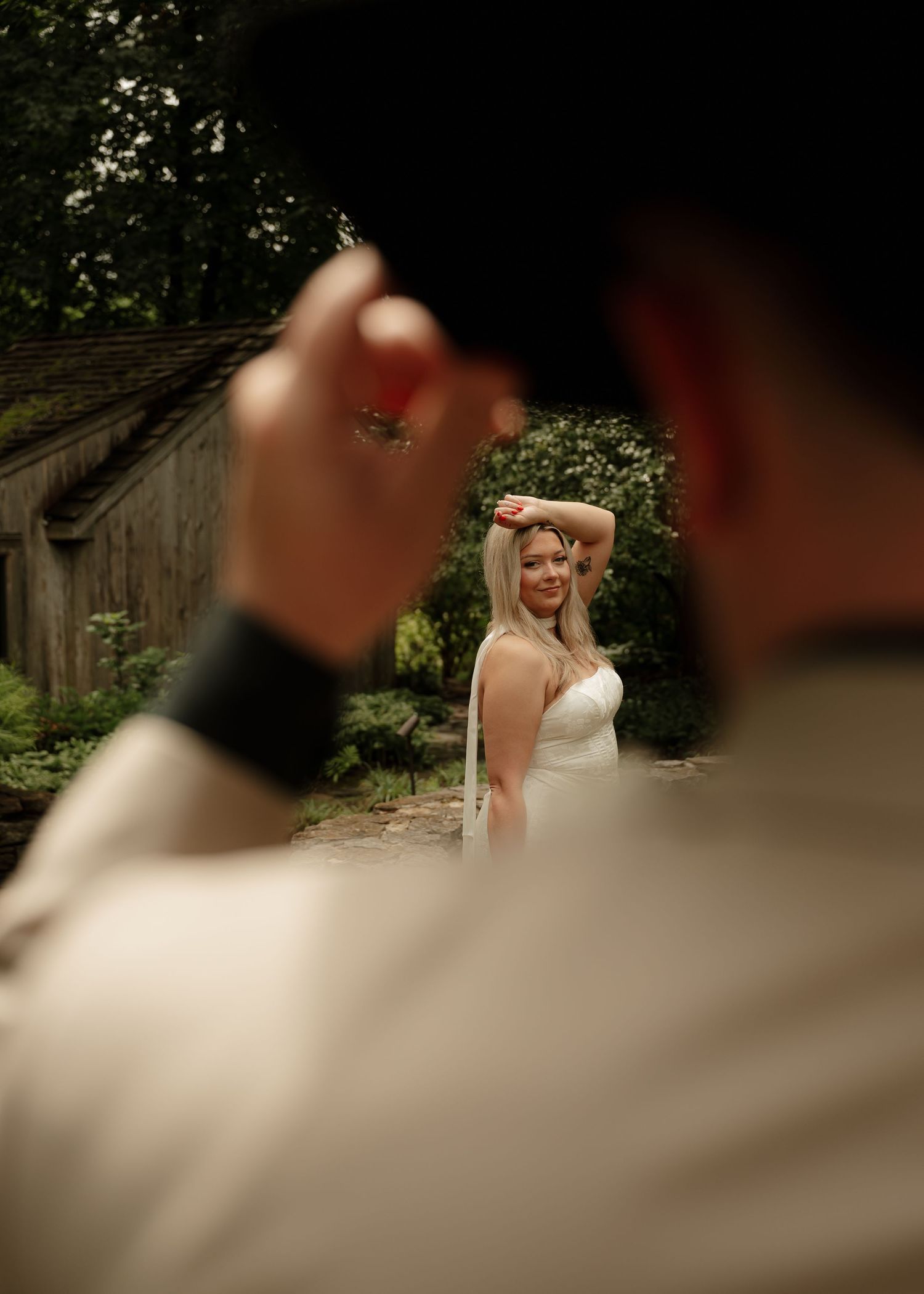 Bride in white dress poses outdoors near rustic barn while groom in foreground raises hand during wedding photo session.