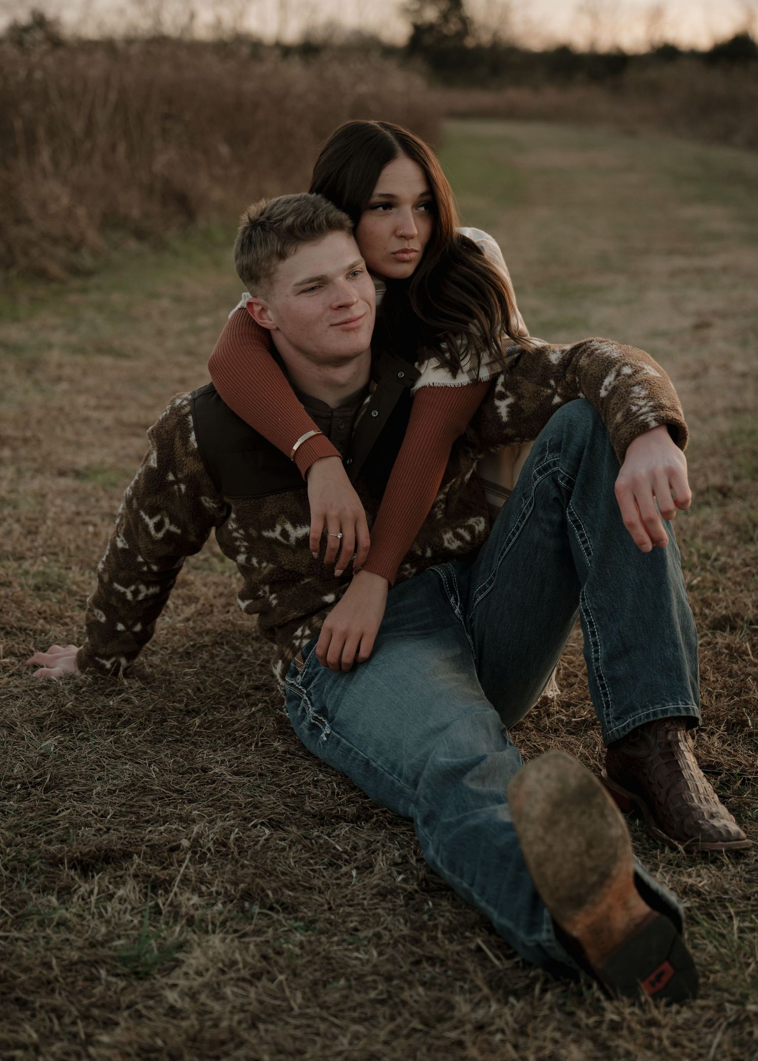 Couple sitting together in a golden field at sunset, woman embracing man from behind in cozy autumn attire.
