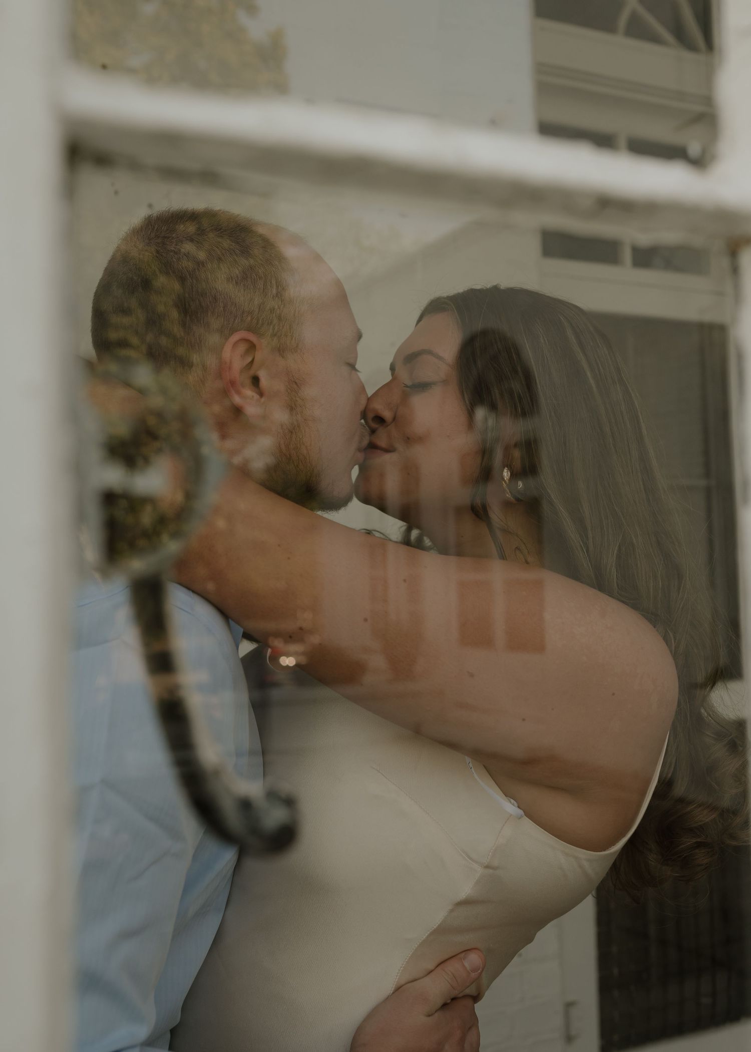 Bride and groom sharing a romantic kiss through a window reflection on their wedding day.