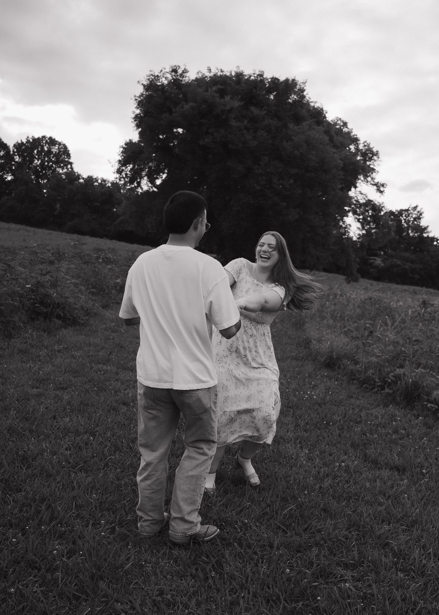 Black and white photo of a couple dancing and laughing together in a grassy field with trees in the background.